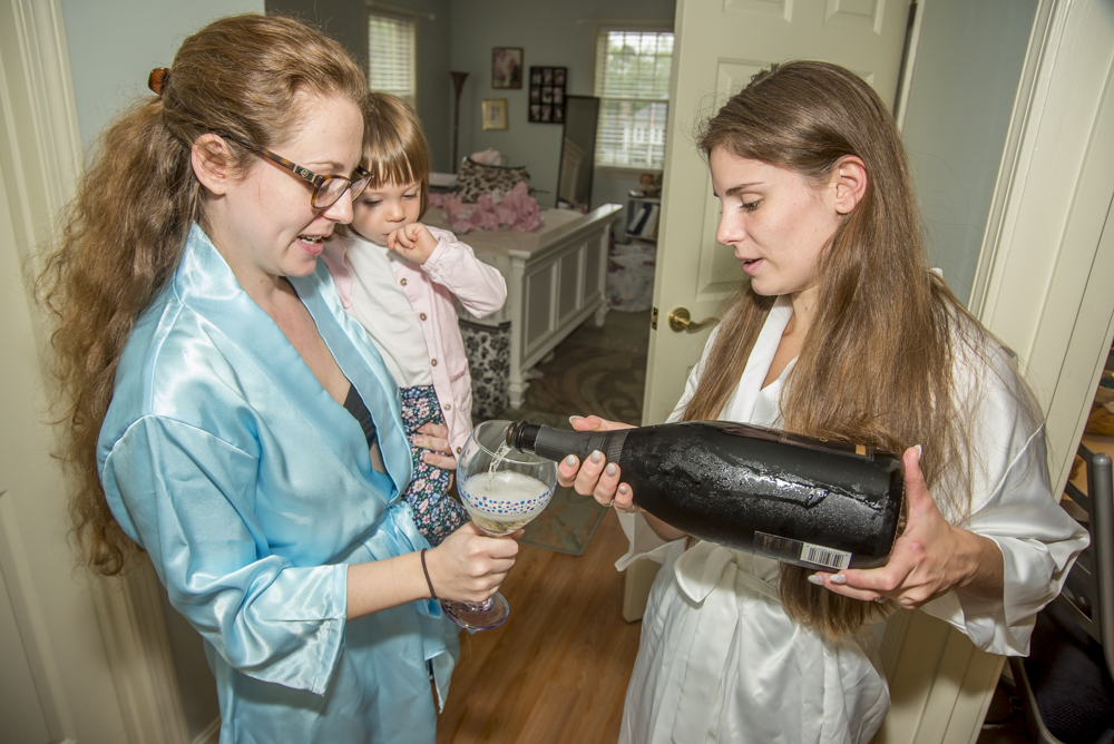 A woman in a blue satin robe pours sparkling wine into a glass held by another woman in a white robe, with a young girl observing in the background.