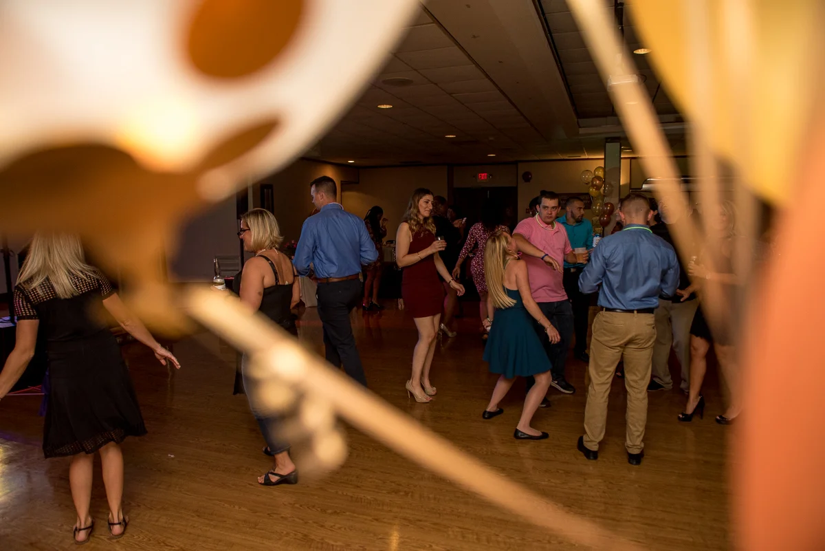 People dancing and socializing at a party or dance event in a room decorated with balloons, viewed through a blurry foreground object.