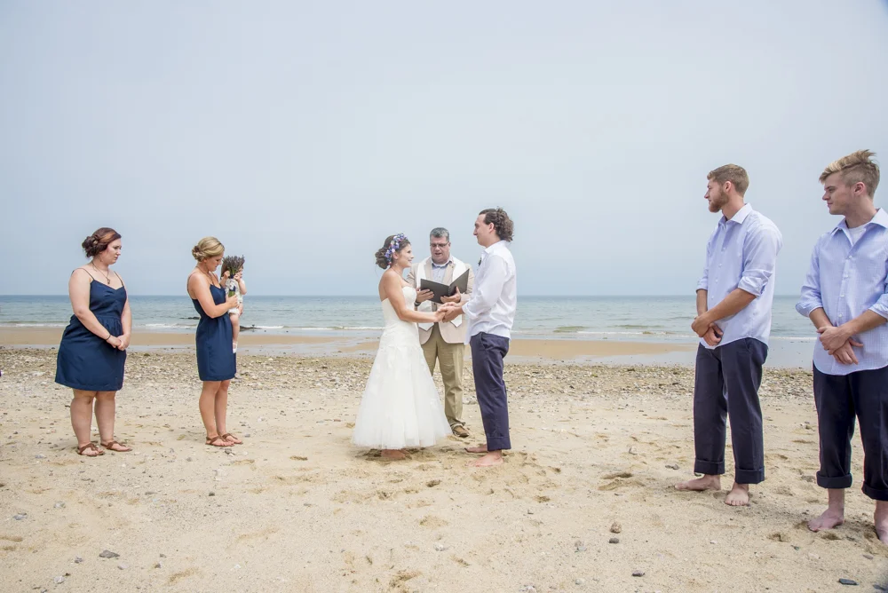 A beach wedding ceremony with a couple exchanging vows, officiant, bridesmaids in blue, groomsmen in light shirts, and ocean in the background.