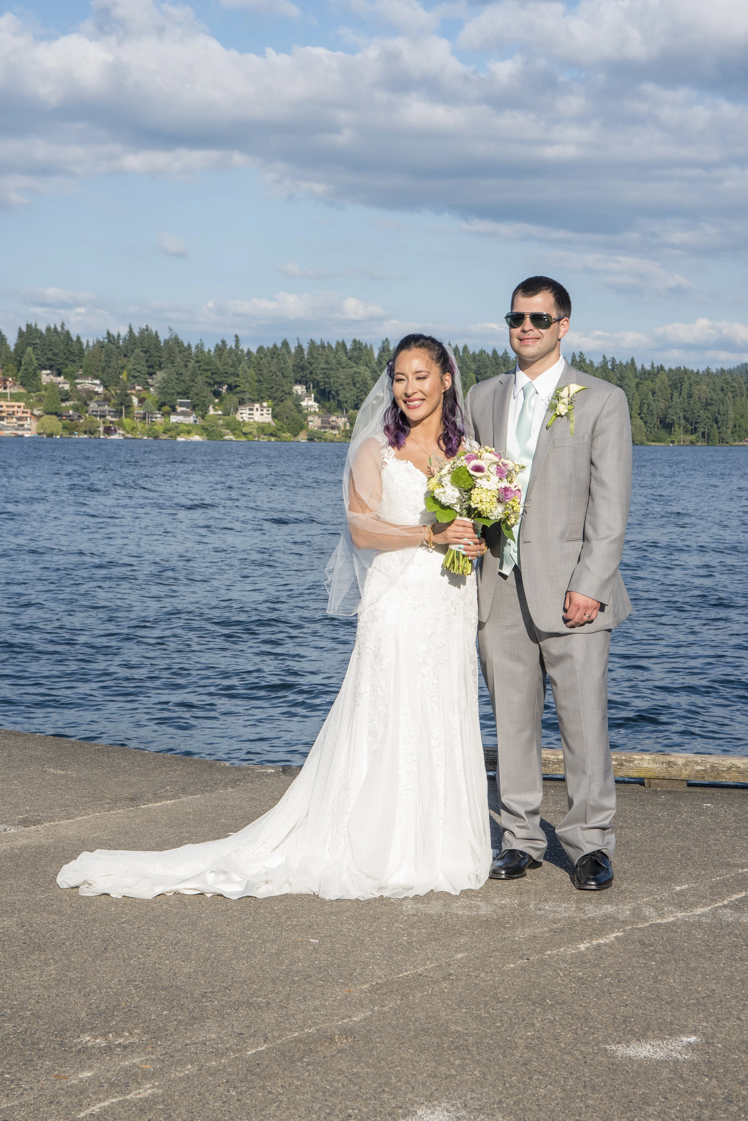 A newlywed couple standing on a dock by a lake, smiling. The bride is wearing a white wedding dress with a veil and holding a bouquet, while the groom is dressed in a light gray suit with a boutonniere and sunglasses. There are trees and houses acros