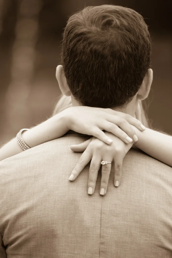 A woman with her arms wrapped around a man's neck, visible from the back, with her hands resting on his shoulders. The woman is wearing a bracelet and a ring on her finger.