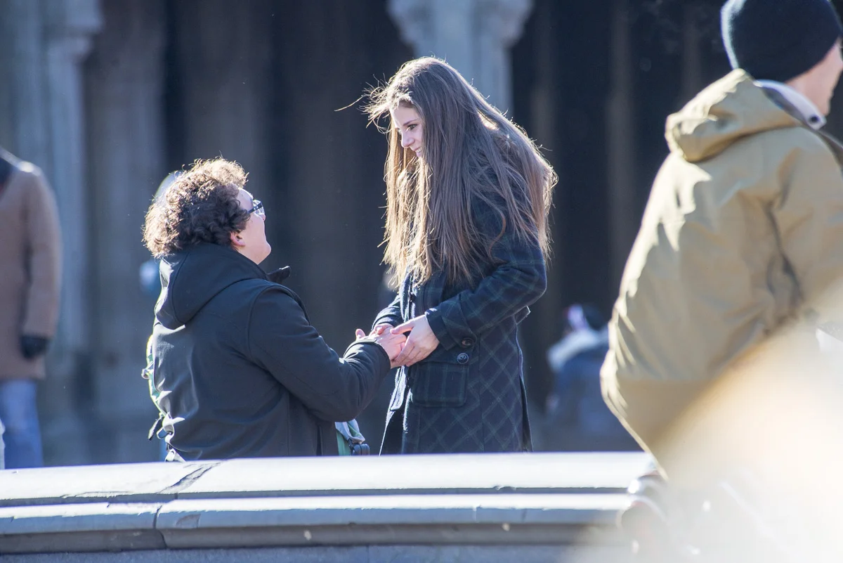 A young woman is kneeling and holding hands with a man, smiling at each other outdoors on a bright day, with several people in the background.