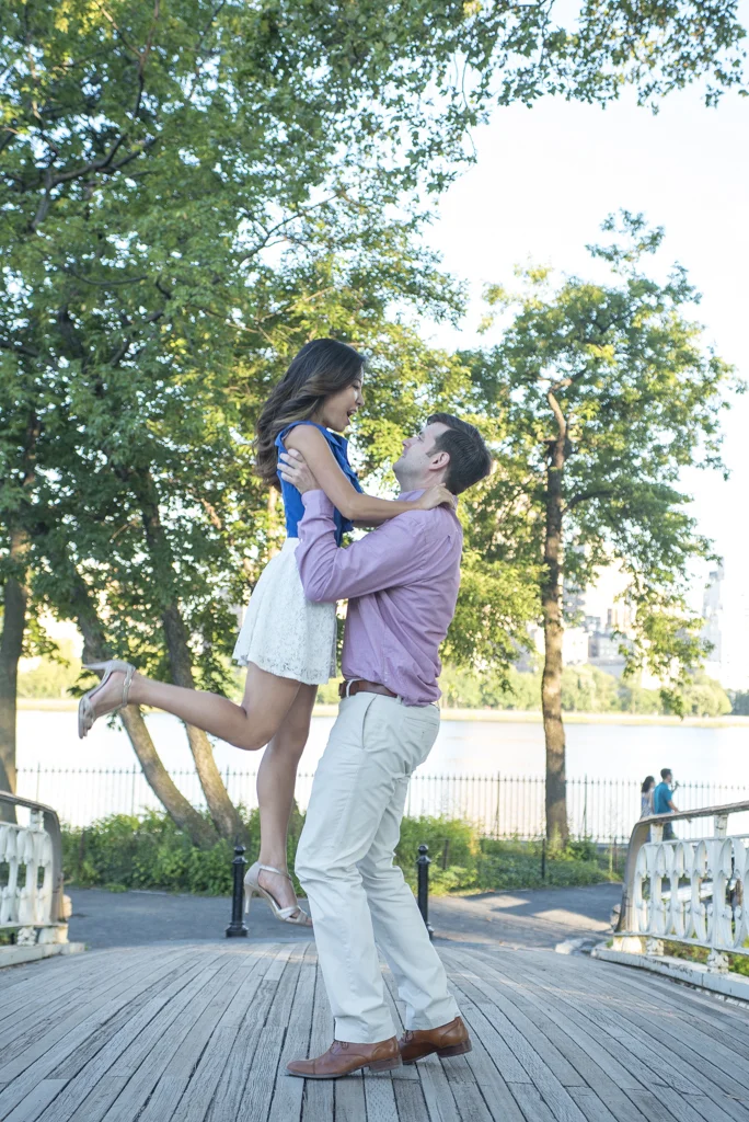 A man lifting a woman in a park by a river, surrounded by trees, during daytime.