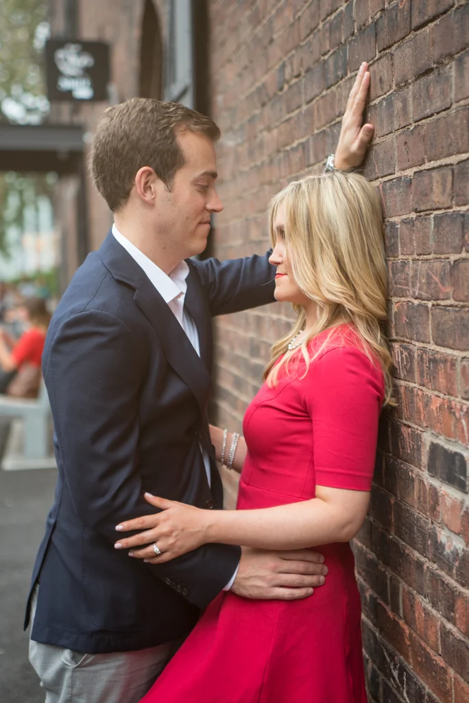 A man and woman in formal attire, standing close against a brick wall. The man is touching the wall with one hand, while the woman has her hand on his waist. They are gazing at each other romantically.