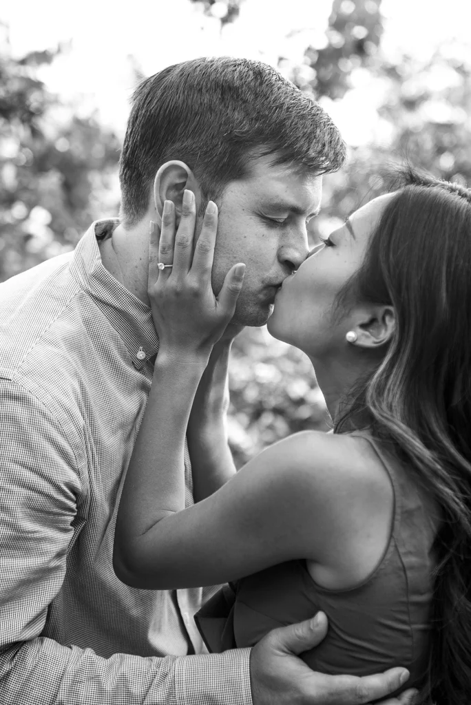 A black-and-white photo of a couple kissing outdoors, with the woman gently holding the man's face and her arm around his shoulder.