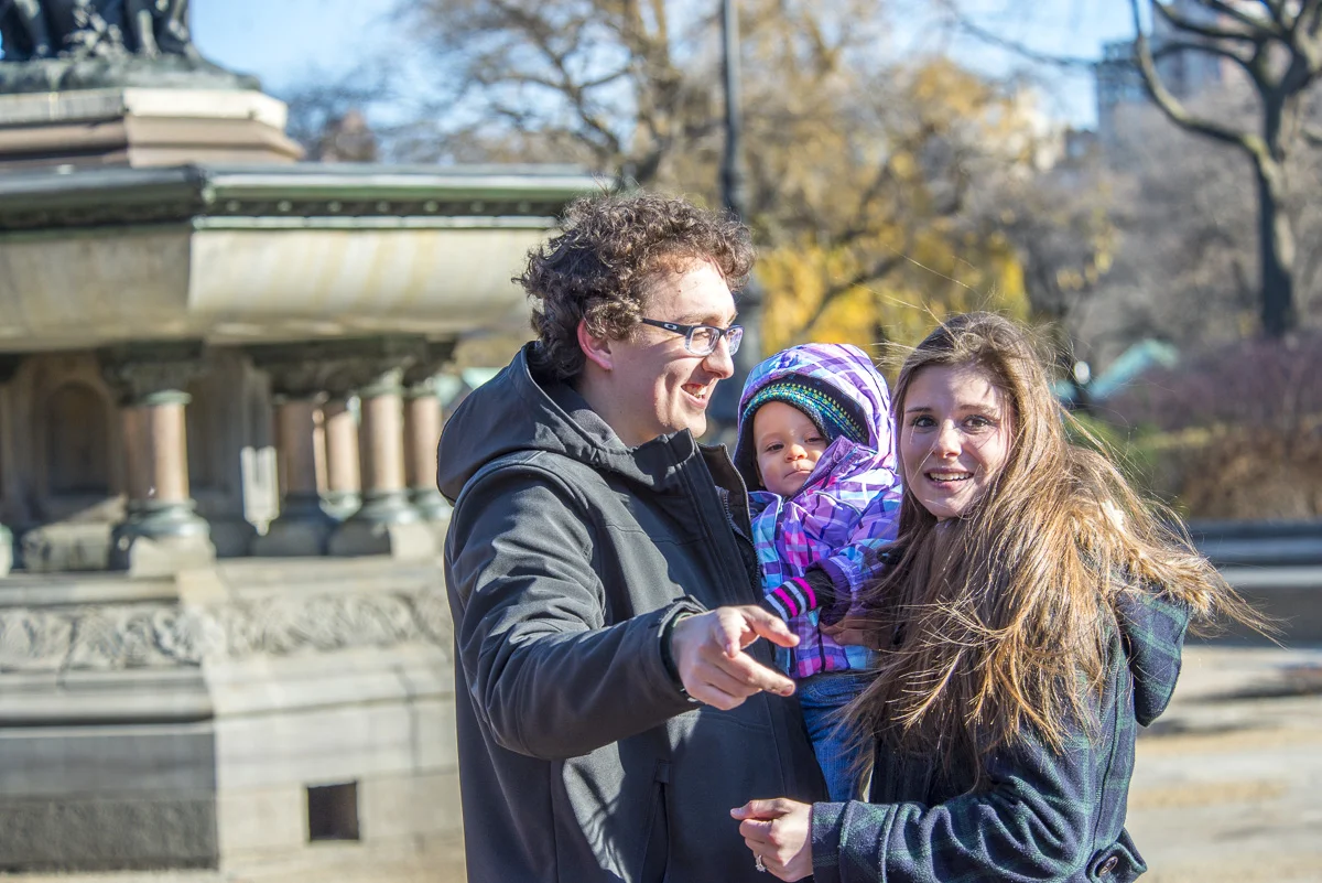 A man, woman, and a small child are outdoors in a park with trees and a fountain in the background. The man is holding the child and smiling, the woman is standing close and smiling, and the child is looking at the camera.