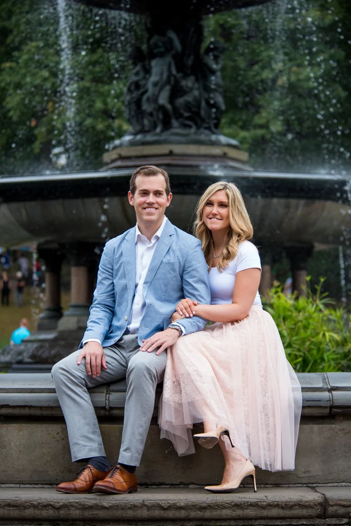 A couple sitting on a stone bench in front of a fountain in a park, smiling. The man is wearing a light blue blazer and gray pants; the woman is wearing a white top and a long, light-colored skirt.