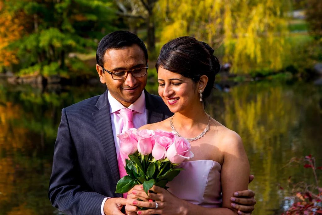 A couple, the woman in a strapless pink dress and the man in a suit with a pink tie, standing outdoors by a lake with autumn trees, holding a bouquet of pink roses.