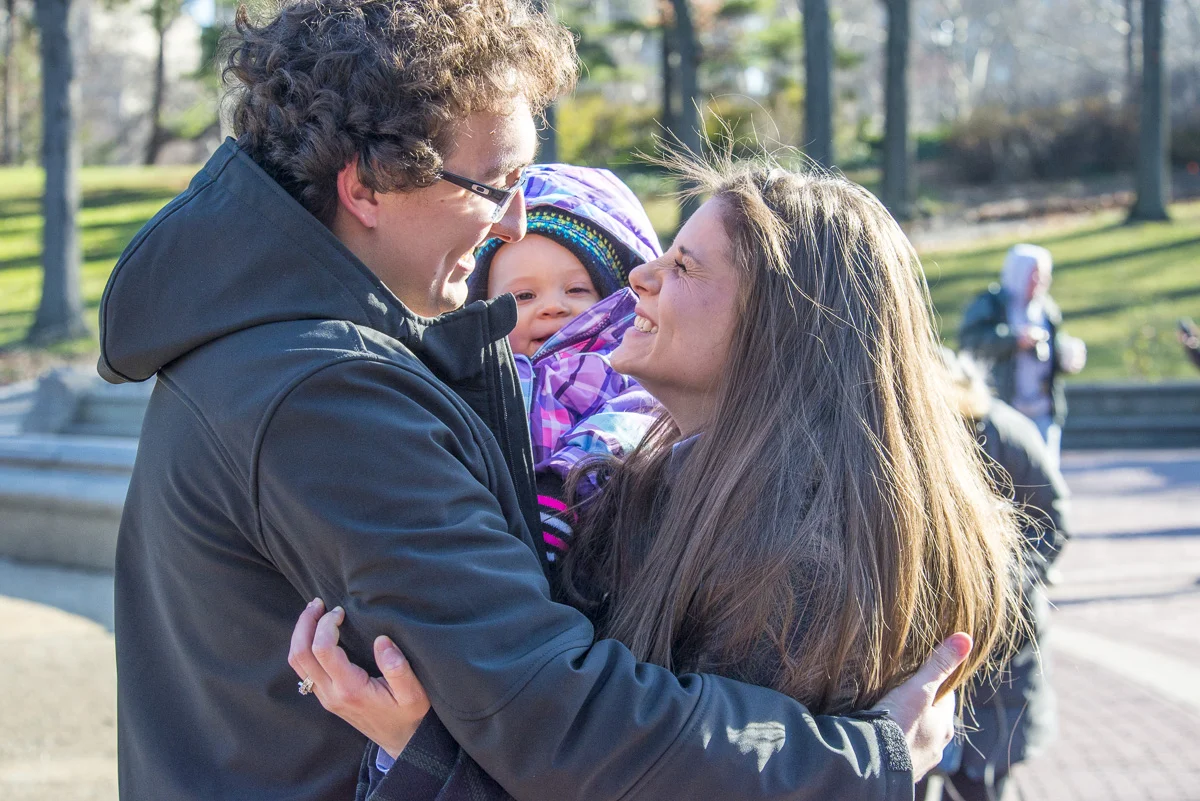 A family of three, a man, woman, and a young girl, sharing a joyful embrace outdoors in a park.