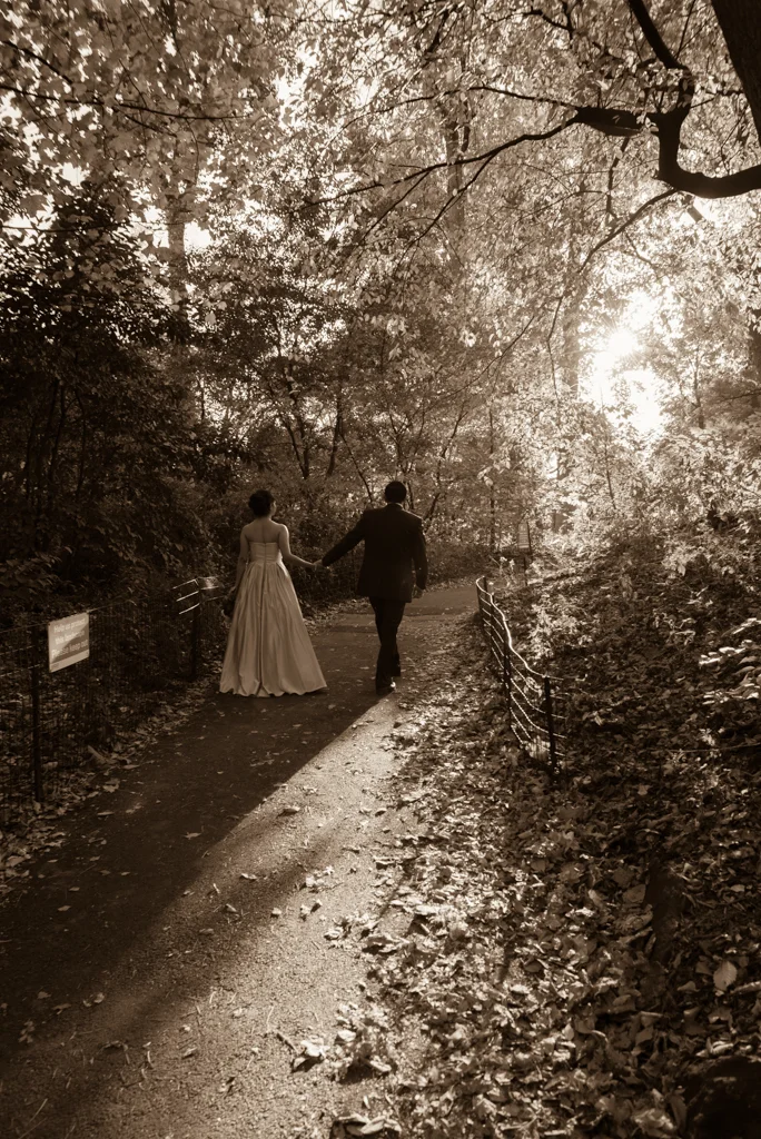 A couple walking hand in hand on a forest trail during sunset, with trees and fallen leaves surrounding them.