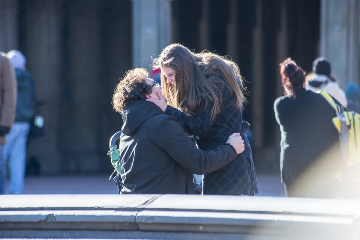Two young women are hugging and touching faces, with other people in the background.