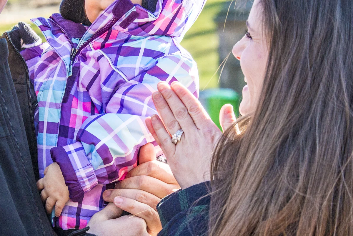 A woman with a wedding ring on her finger praying with her hands clasped in front of a child wearing a purple and pink patterned winter jacket outdoors.