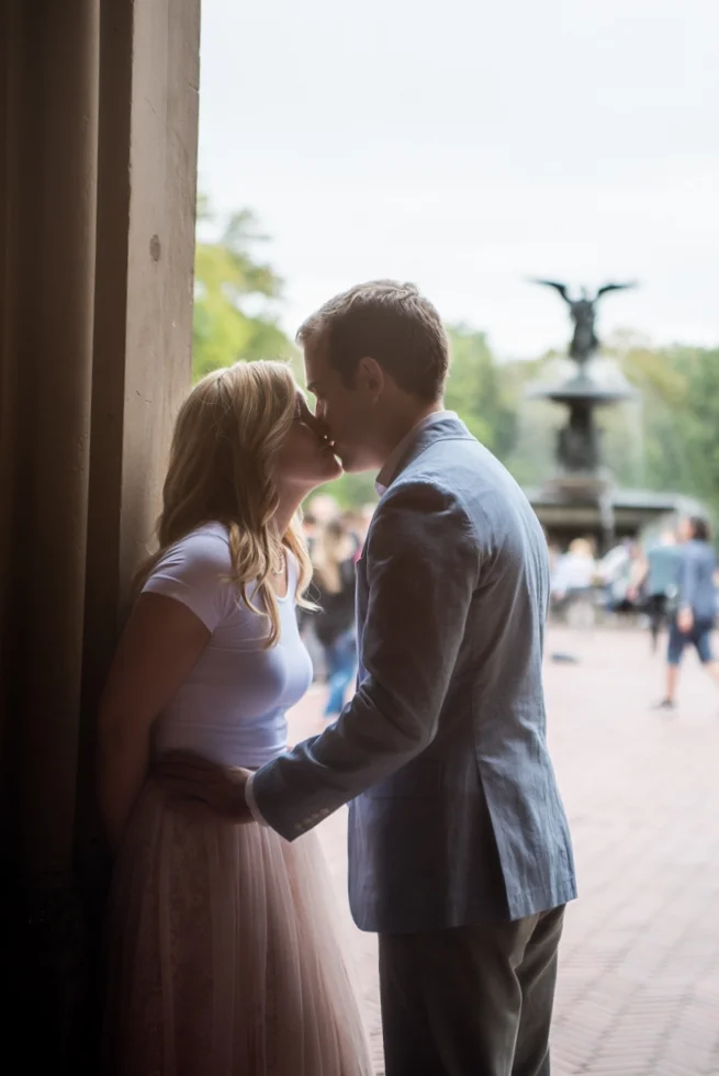 A couple sharing a kiss, standing close near a wooden wall, with a fountain and outdoor scene in the background.
