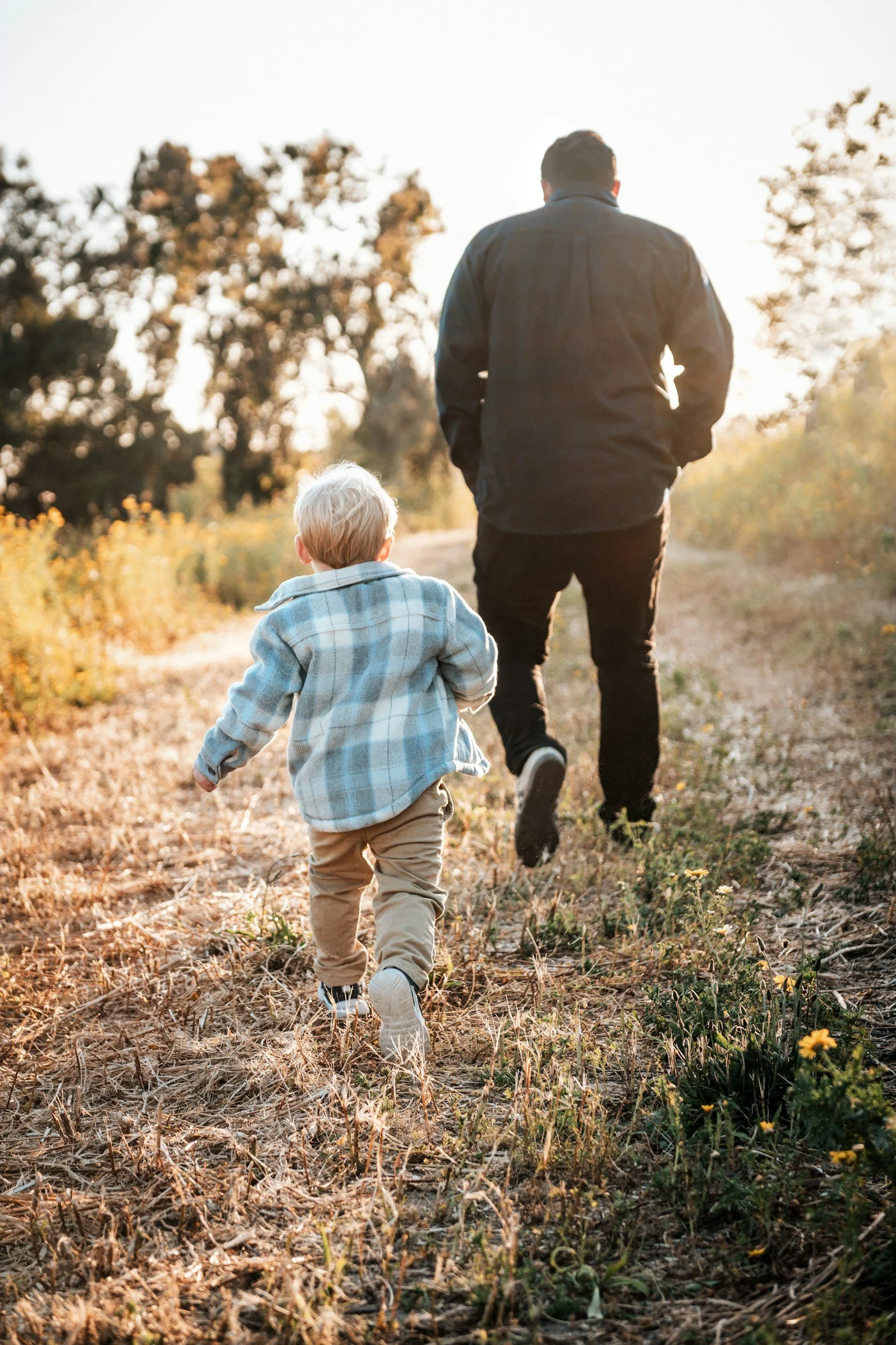 A man and a young child walk together on a dirt path through a field lined with trees and yellow flowers during sunset.