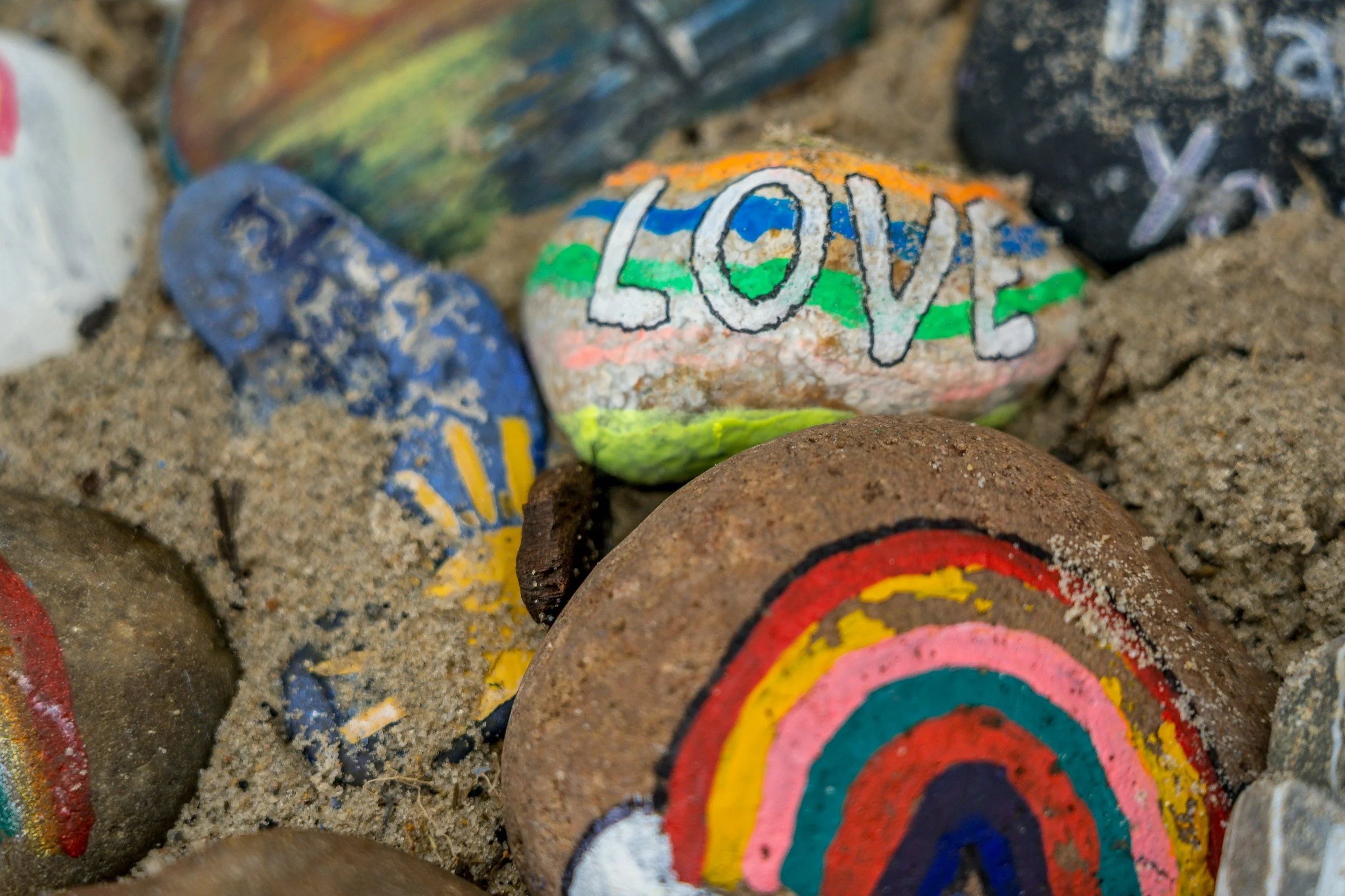 Painted rocks with rainbow and the word 'LOVE' on one of them, placed on sandy ground.