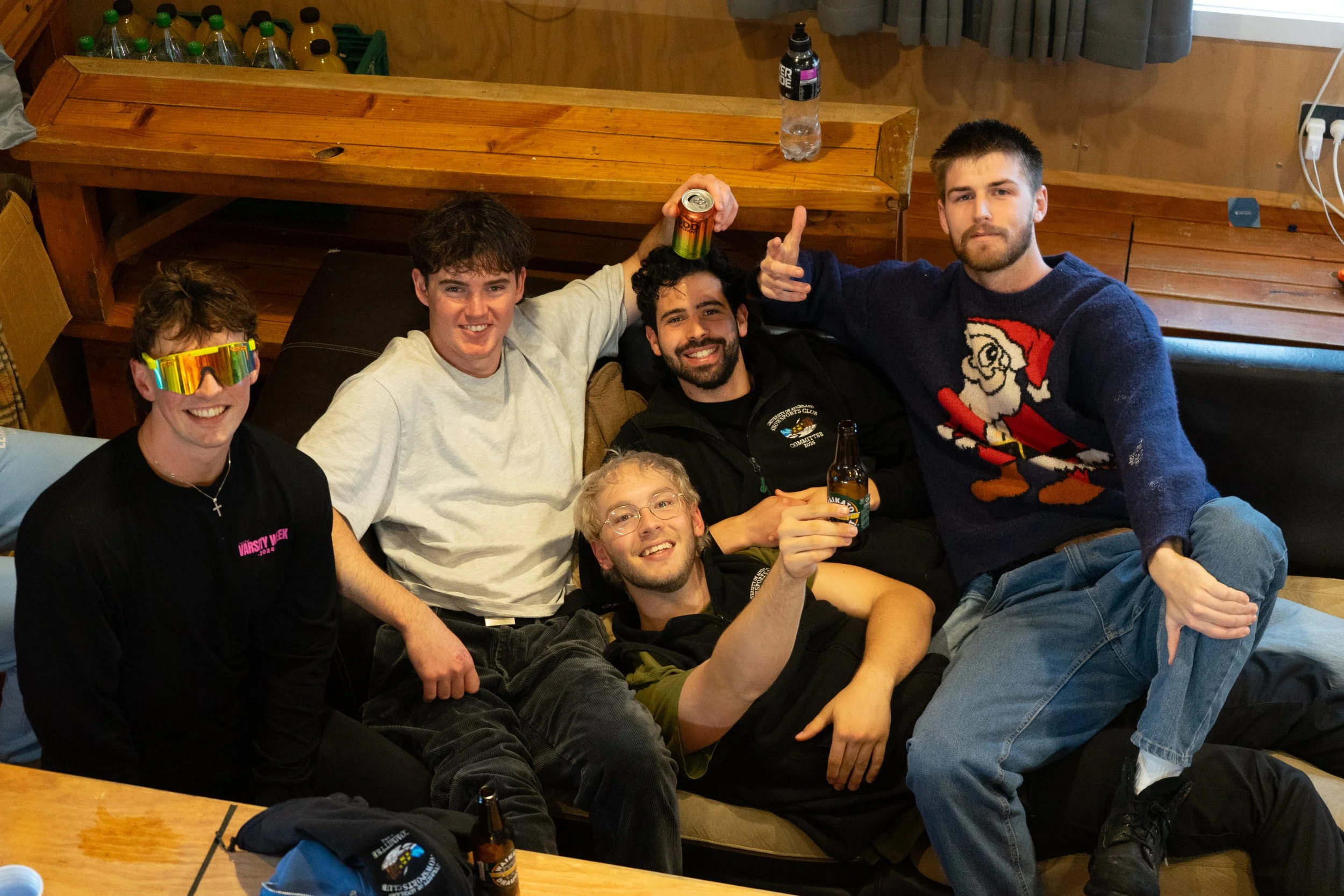 Five young men sitting on a couch in a casual indoor setting, smiling, holding drinks, with wood-paneled walls, a wooden table, and bottles of water and soda in the background.