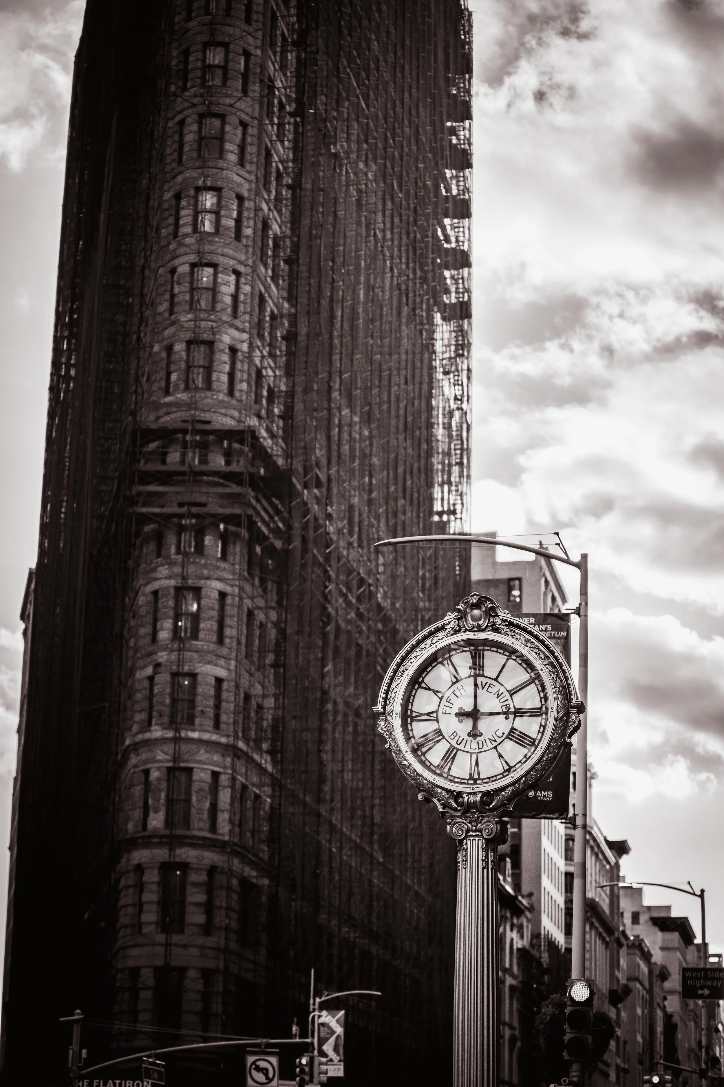 Black and white photo of a city street with a vintage clock showing 2 o'clock and a tall building under construction in the background.