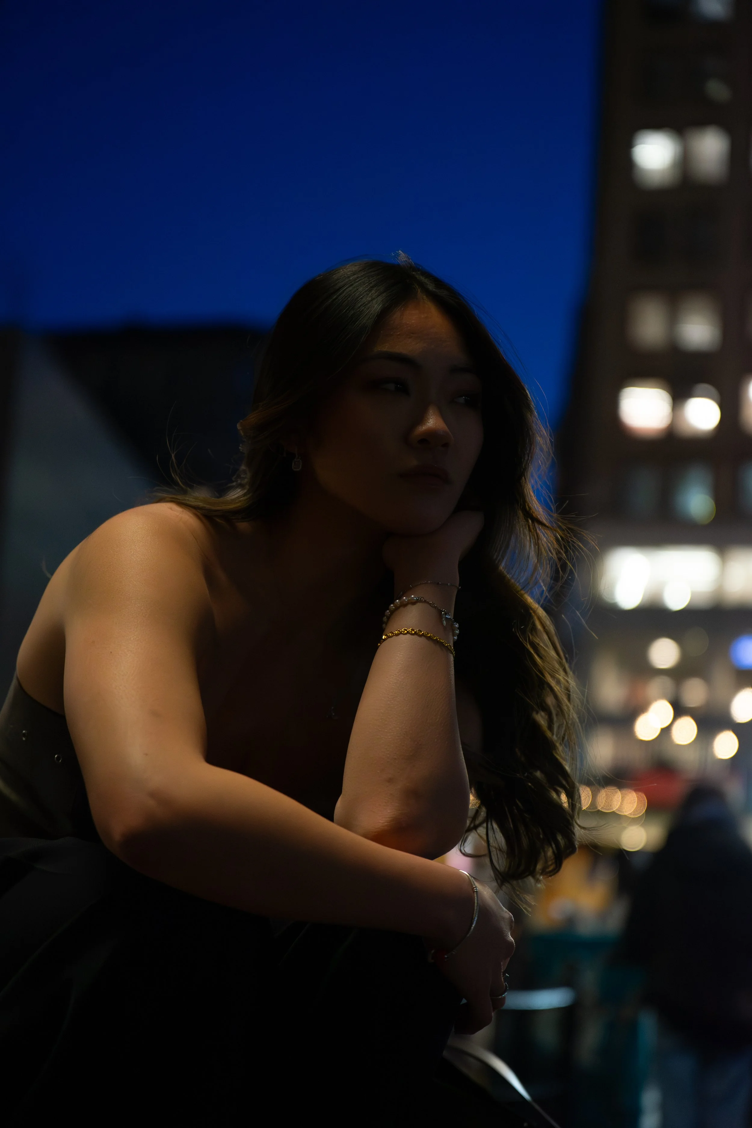 A woman with long wavy hair, wearing jewelry, sitting at night with her chin resting on her hand, in an outdoor urban setting.