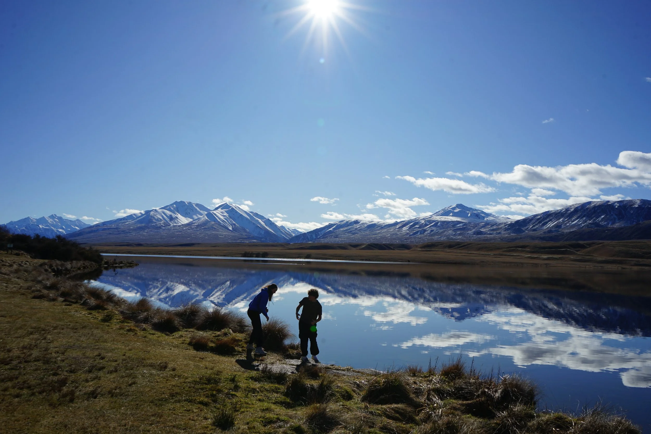 Two children by a lakeside in a mountain landscape with snow-capped peaks, grassy shoreline, and clear blue sky with the sun shining.