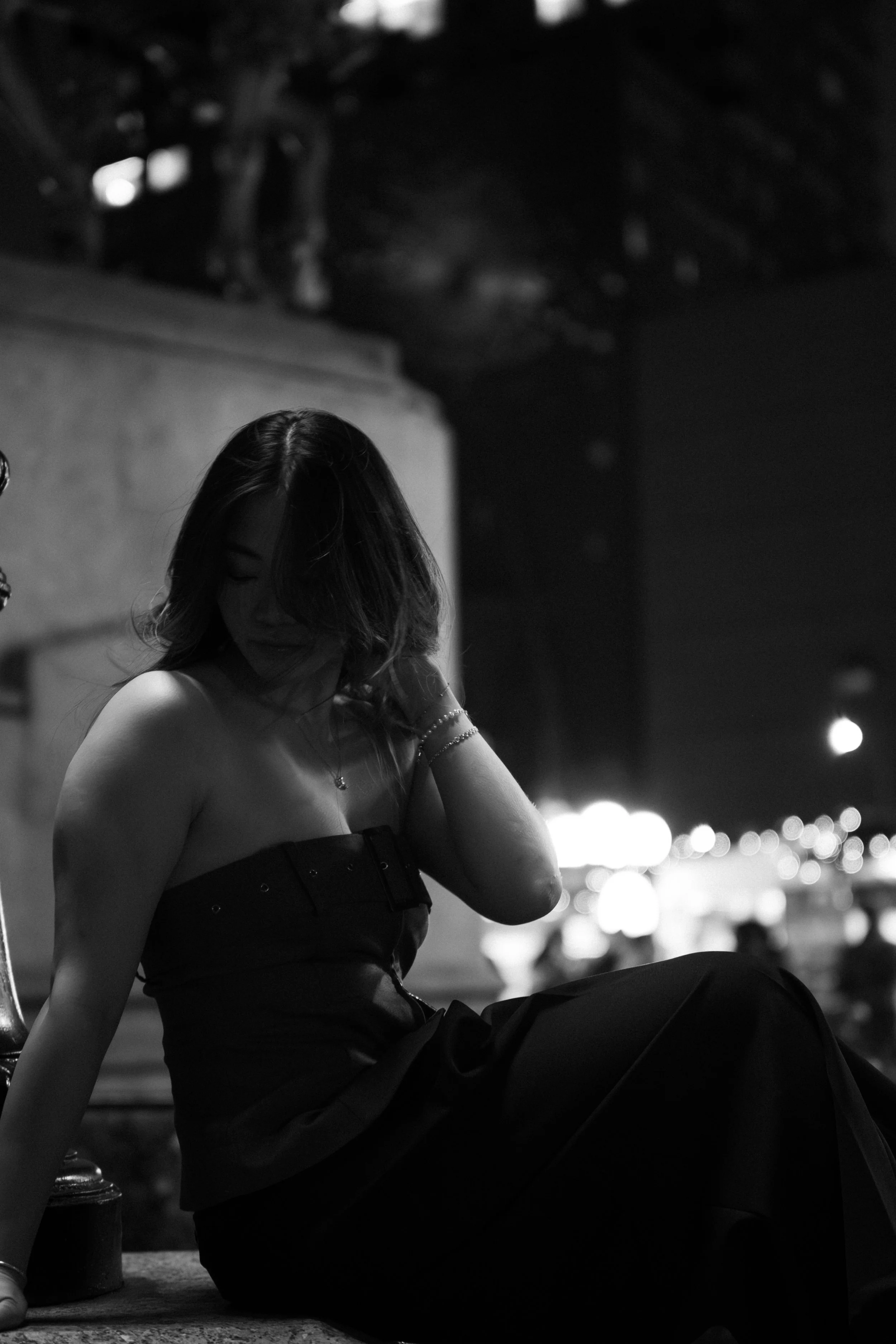 A woman sitting outdoors at night, wearing a strapless dress, with her hair covering part of her face, and city lights in the background.