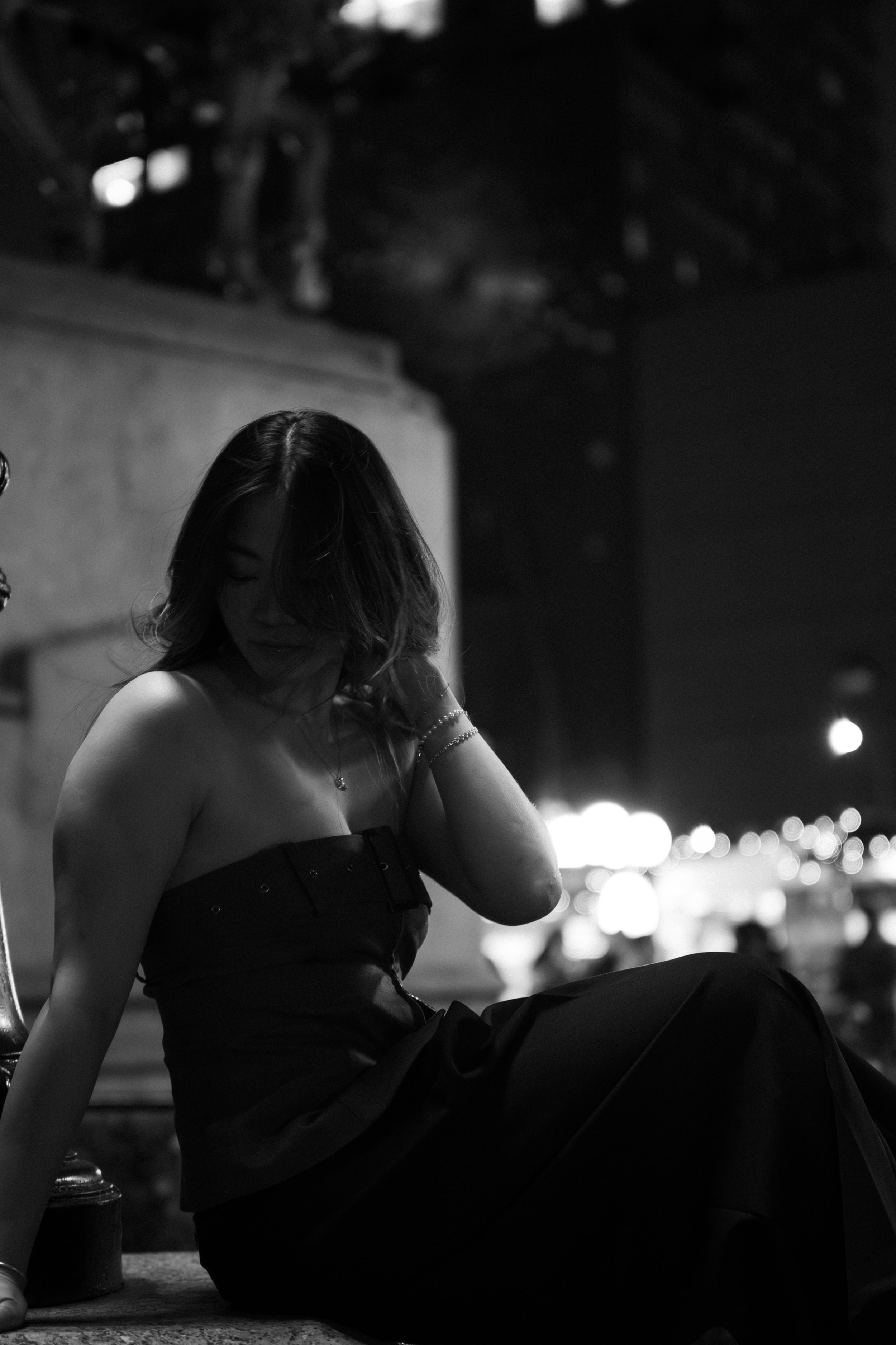 A woman sitting on a ledge at night in a black strapless dress, with her head tilted down and hair covering part of her face, in an urban setting with blurred city lights in the background.