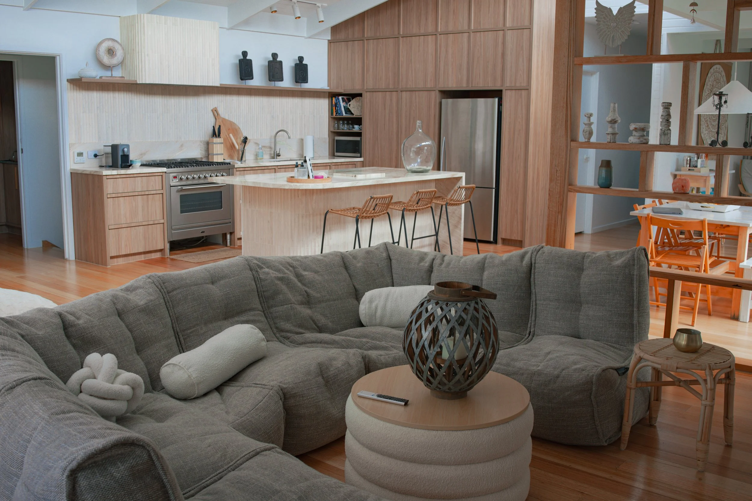 Open-concept living room and kitchen with wooden accents, gray sectional sofa, decorative accessories, and a dining area with orange chairs.