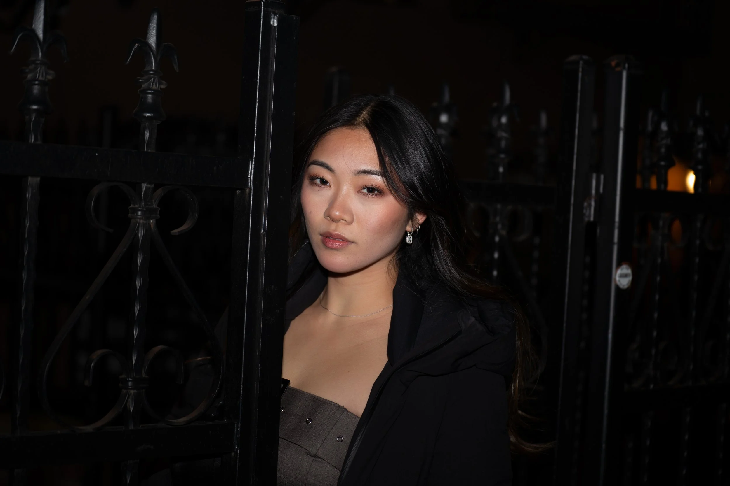 A young woman with dark hair and wearing a black jacket and earrings standing behind a black iron fence at night.