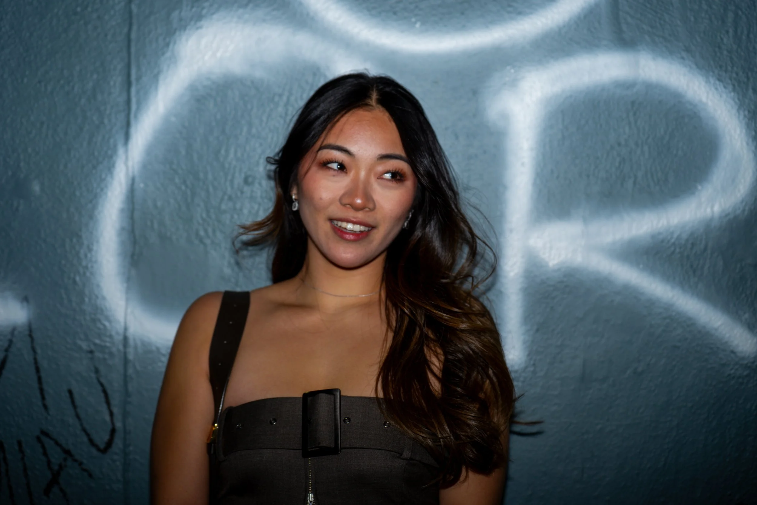 Young woman with long dark hair, wearing a dark sleeveless top, standing in front of a textured blue wall with graffiti and a large white painted shape.
