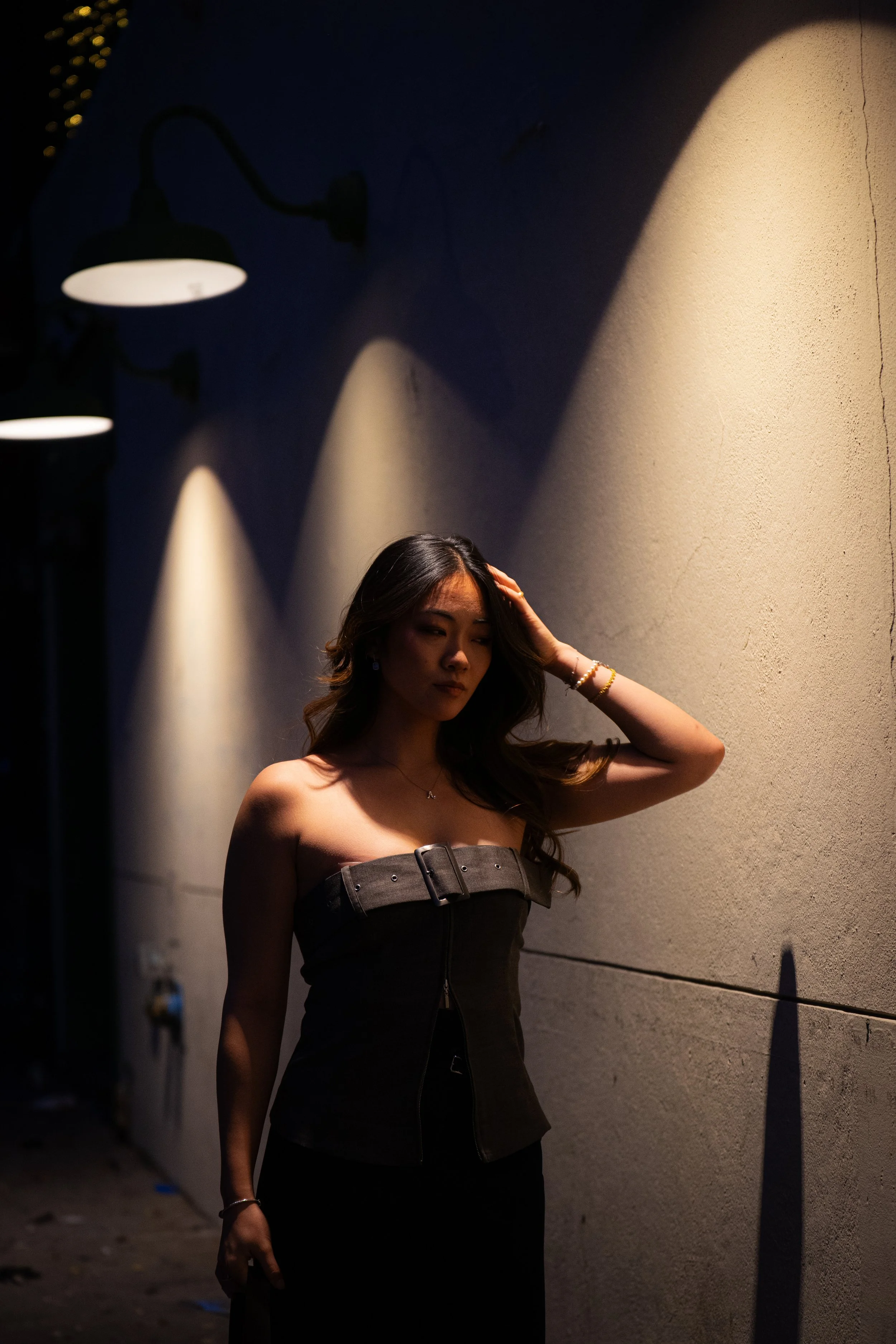 A woman with long dark hair stands in a dimly lit alley at night, with her hand on her head, wearing a strapless black dress. Shadows from overhead lights create patterns on the wall behind her.
