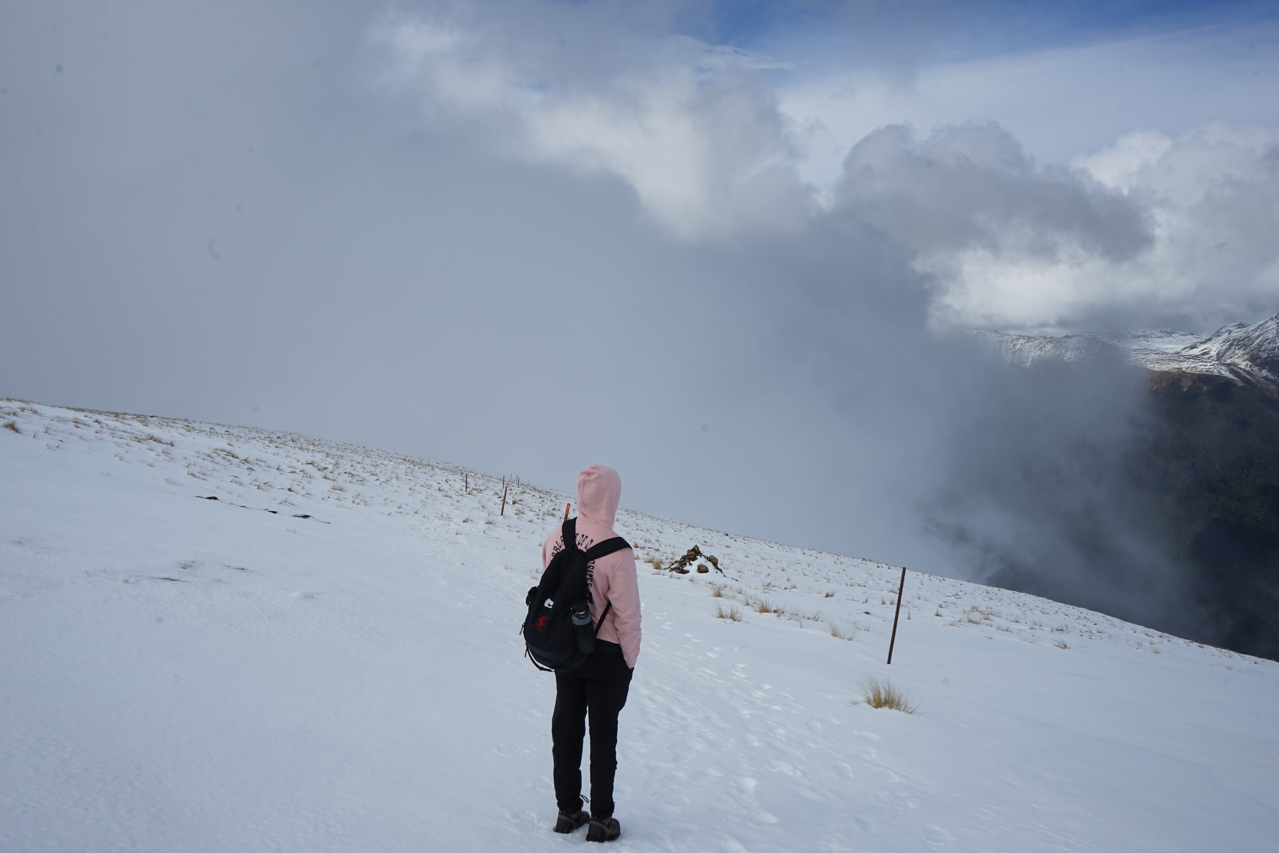 Person wearing a pink hoodie and black pants walking on snowy terrain with a backpack, overlooking a mountain landscape with snow and clouds.
