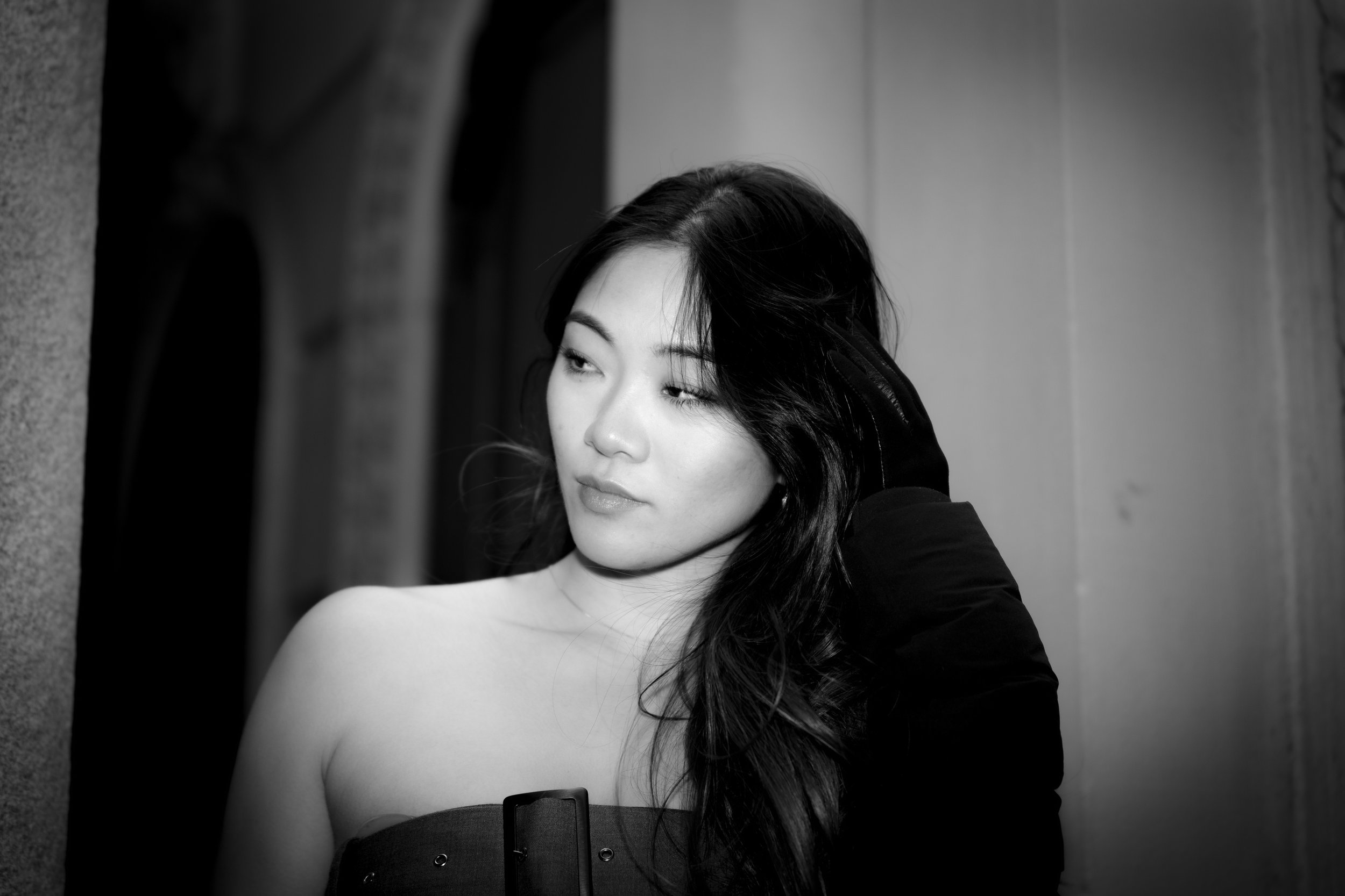 Black and white portrait of a woman with long dark hair, looking to the side, with her hand resting on her head, standing indoors against a plain wall.