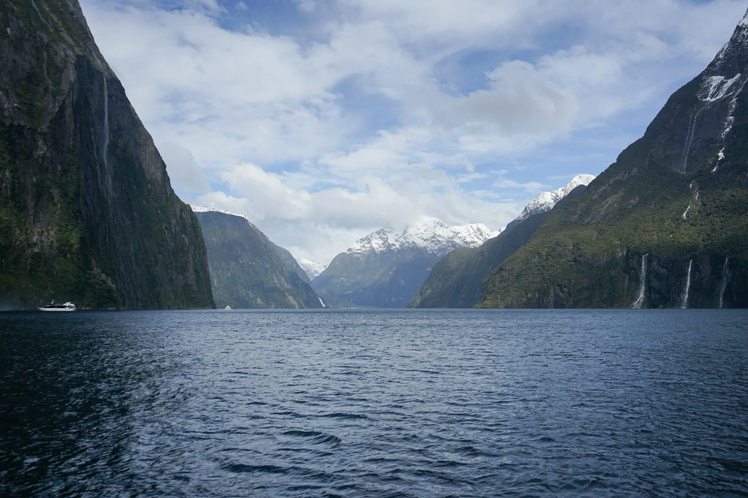 A scenic view of a fjord with tall, steep mountains on each side, some snow-capped, and cascading waterfalls. The water is calm, with gentle ripples, under a partly cloudy sky.