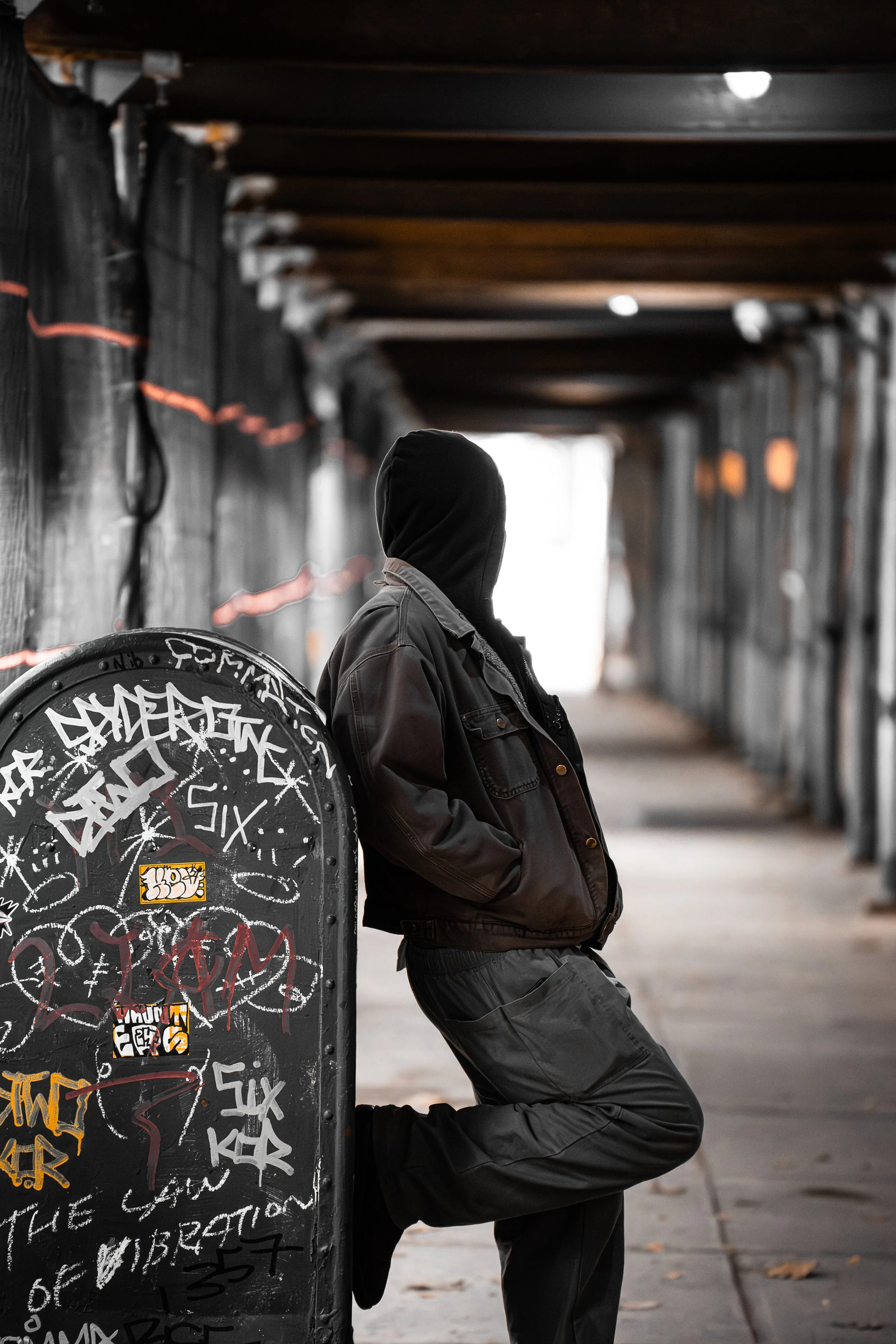 A person in a black hoodie and leather jacket leaning against a graffiti-covered mailbox in a dimly lit urban tunnel.
