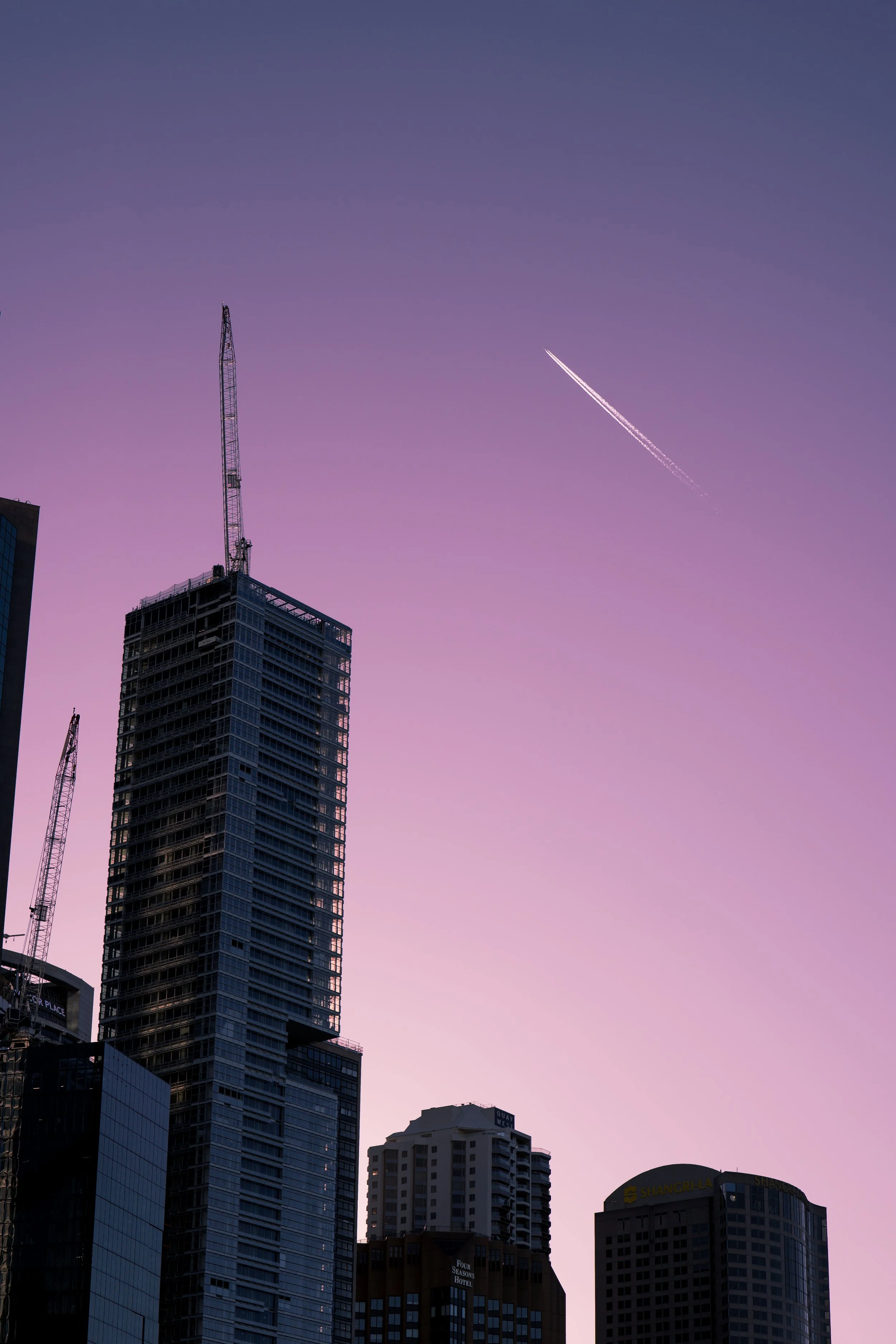 Cityscape featuring high-rise buildings and construction cranes under a purple sky with a jet trail.