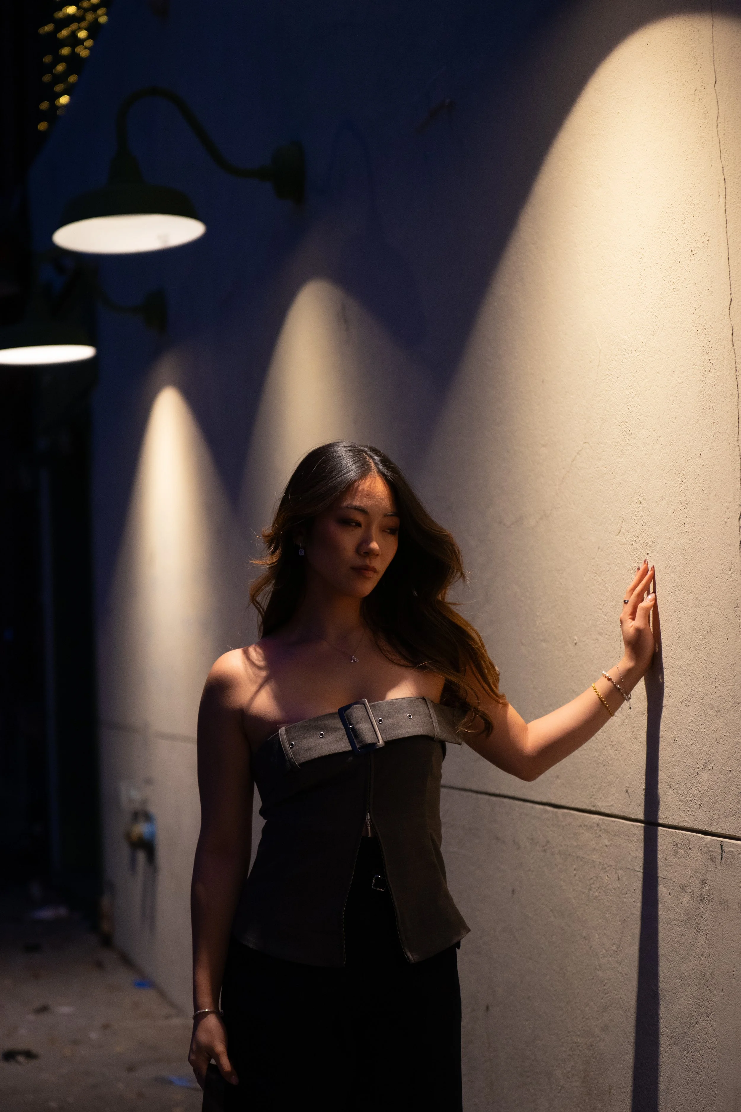 A woman standing against a textured beige wall illuminated by three downward-facing wall-mounted lamps in a dimly lit outdoor setting.