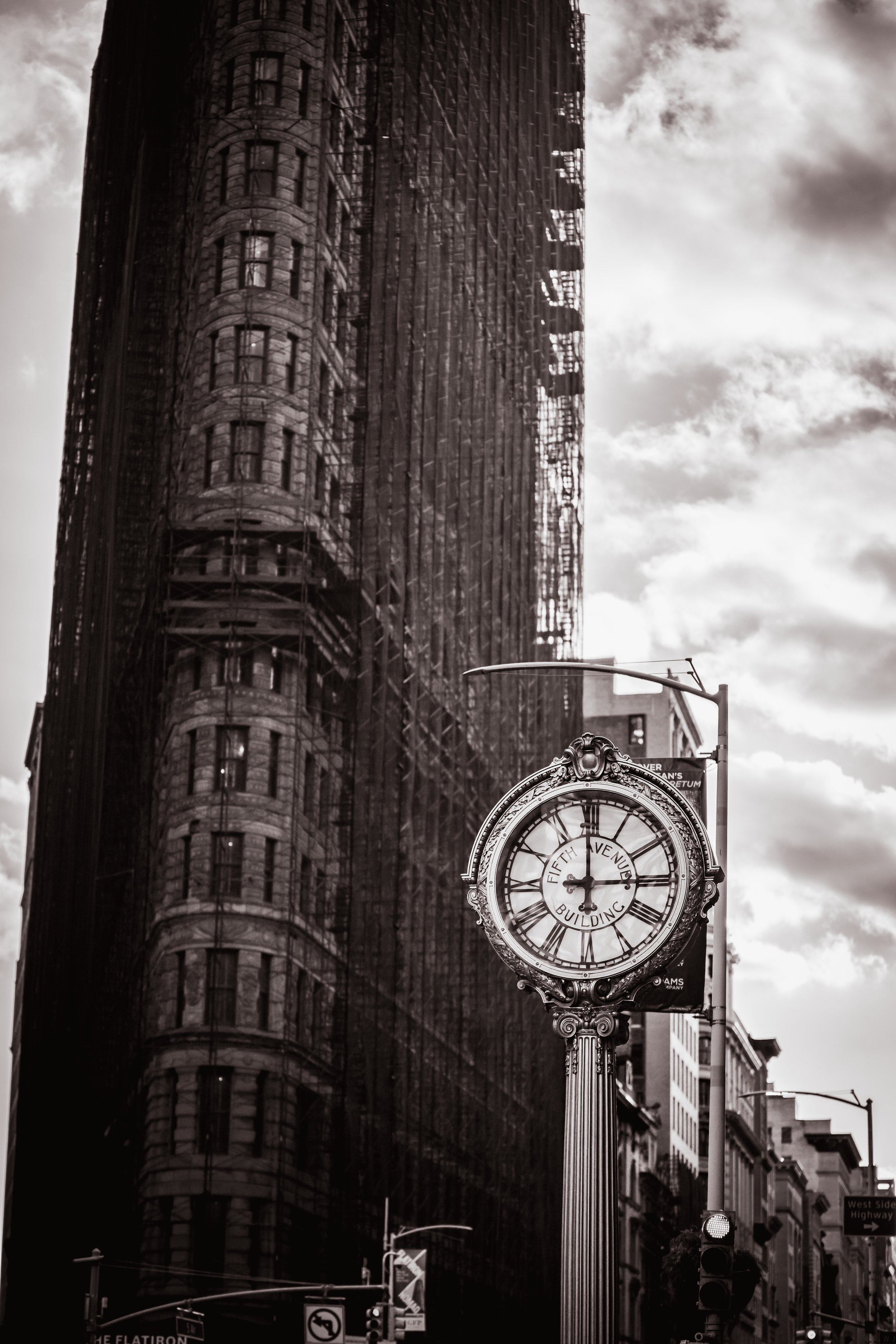 Black and white photo of a street scene with a large clock on a pole, displaying roughly 12:07, against a backdrop of tall buildings with reflective windows and a cloudy sky.