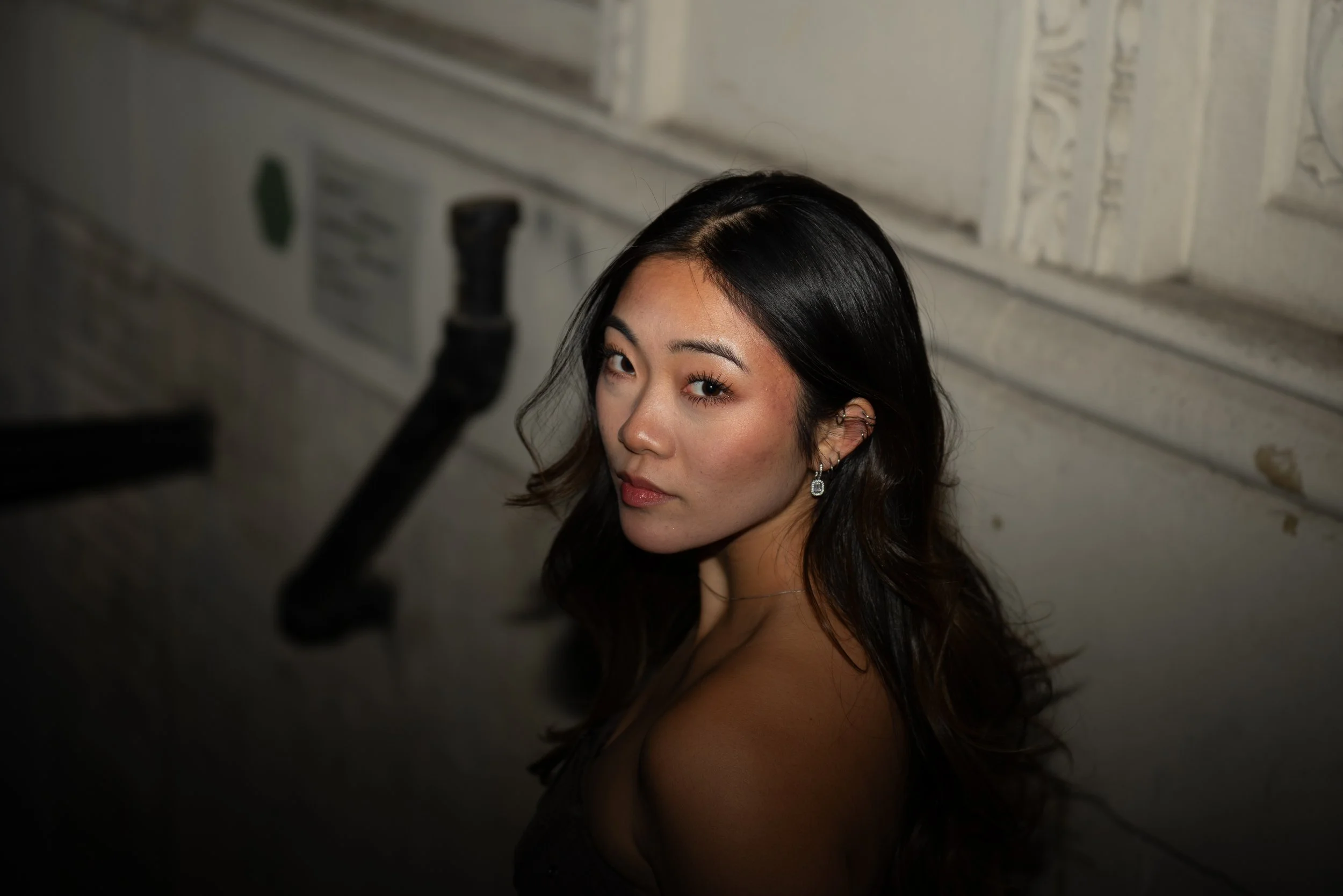 A woman with long dark hair and earrings looking over her shoulder in a dimly lit room with decorative molding on the wall.