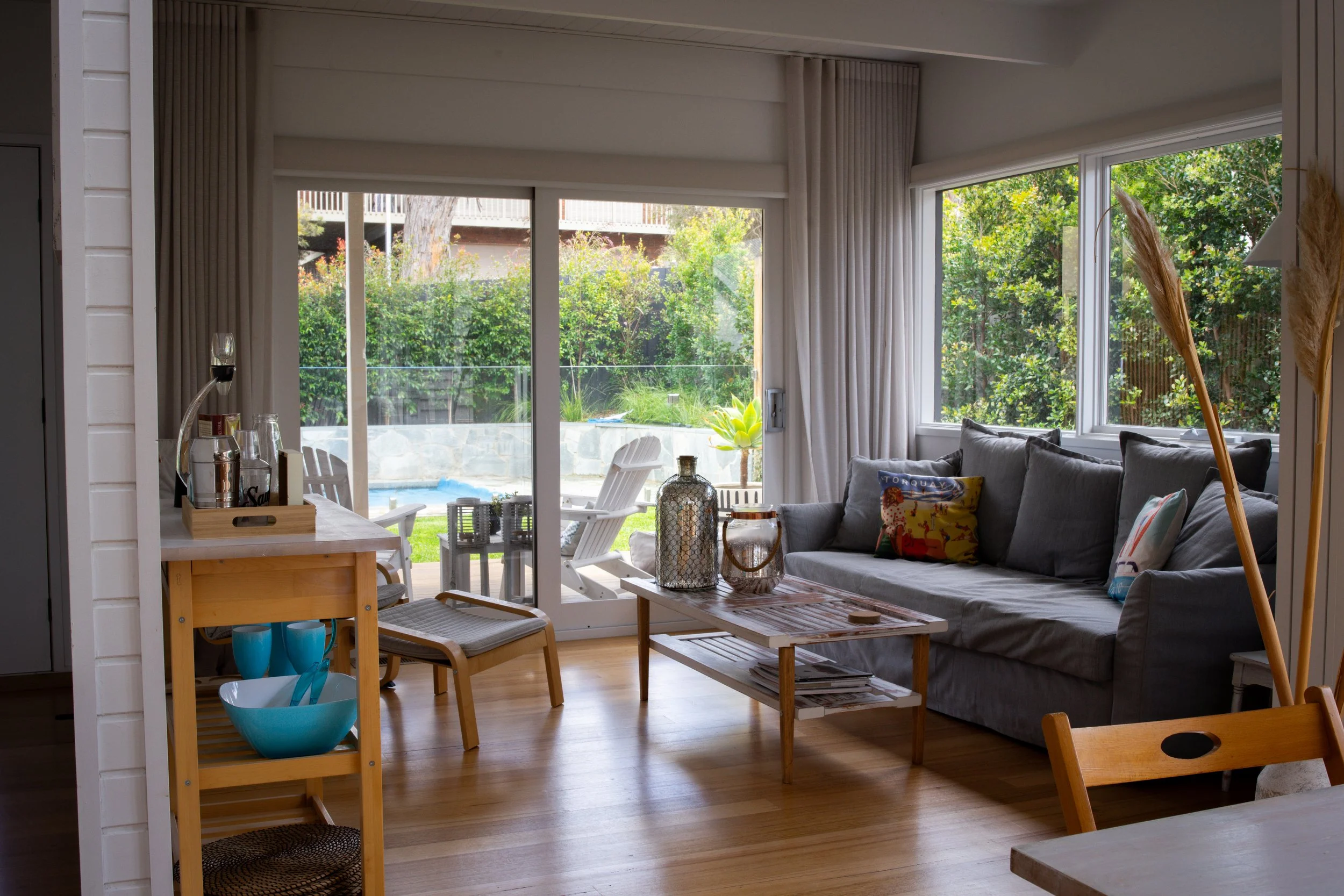 Living room with large windows and sliding glass door, gray sofa with colorful pillows, wooden coffee table with decorative items, and patio chairs outside in a lush backyard.
