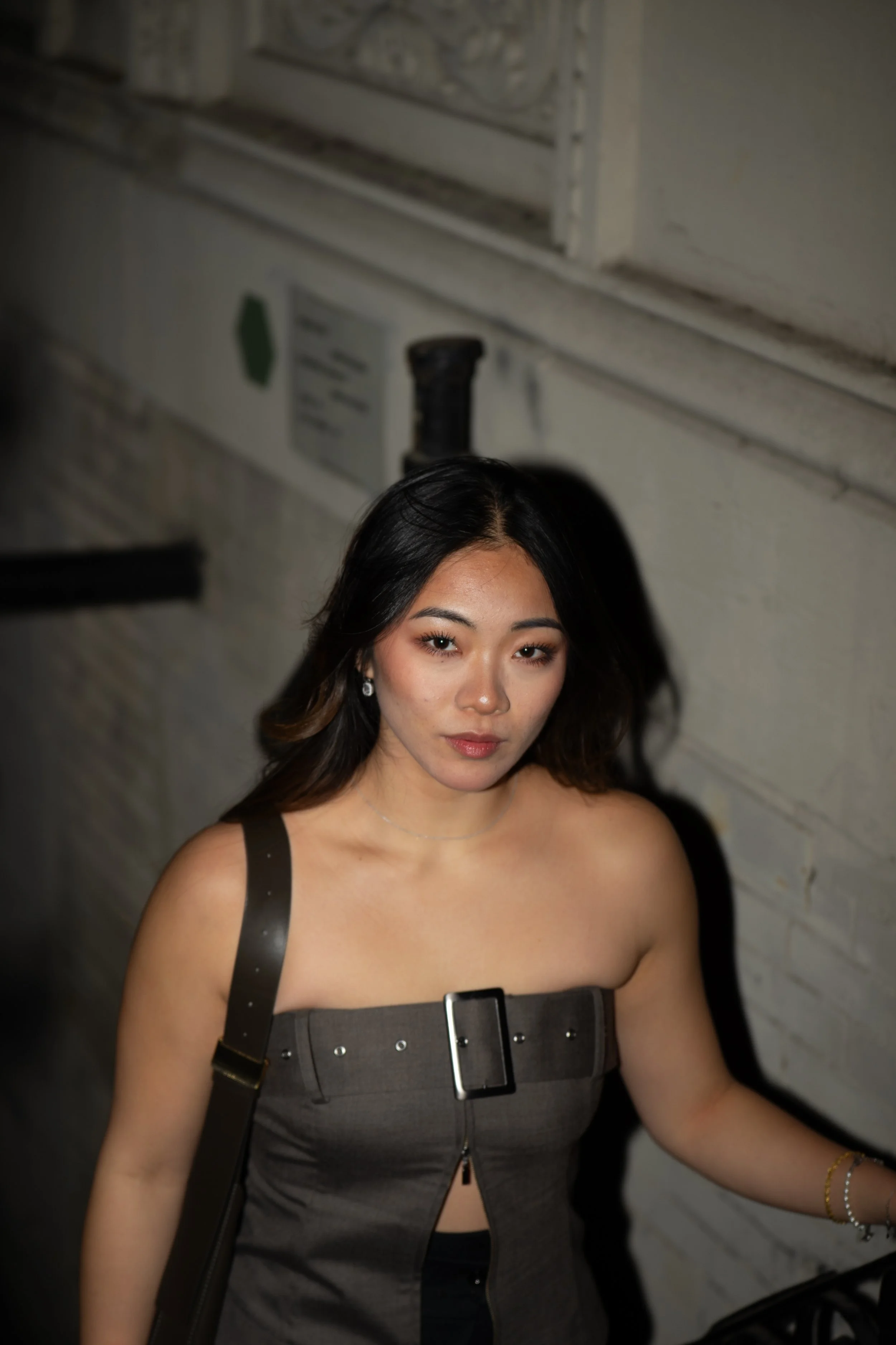 A young woman with dark hair and makeup looking at the camera, wearing a strapless gray dress with a black buckle and carrying a shoulder bag, standing in front of a white brick wall.