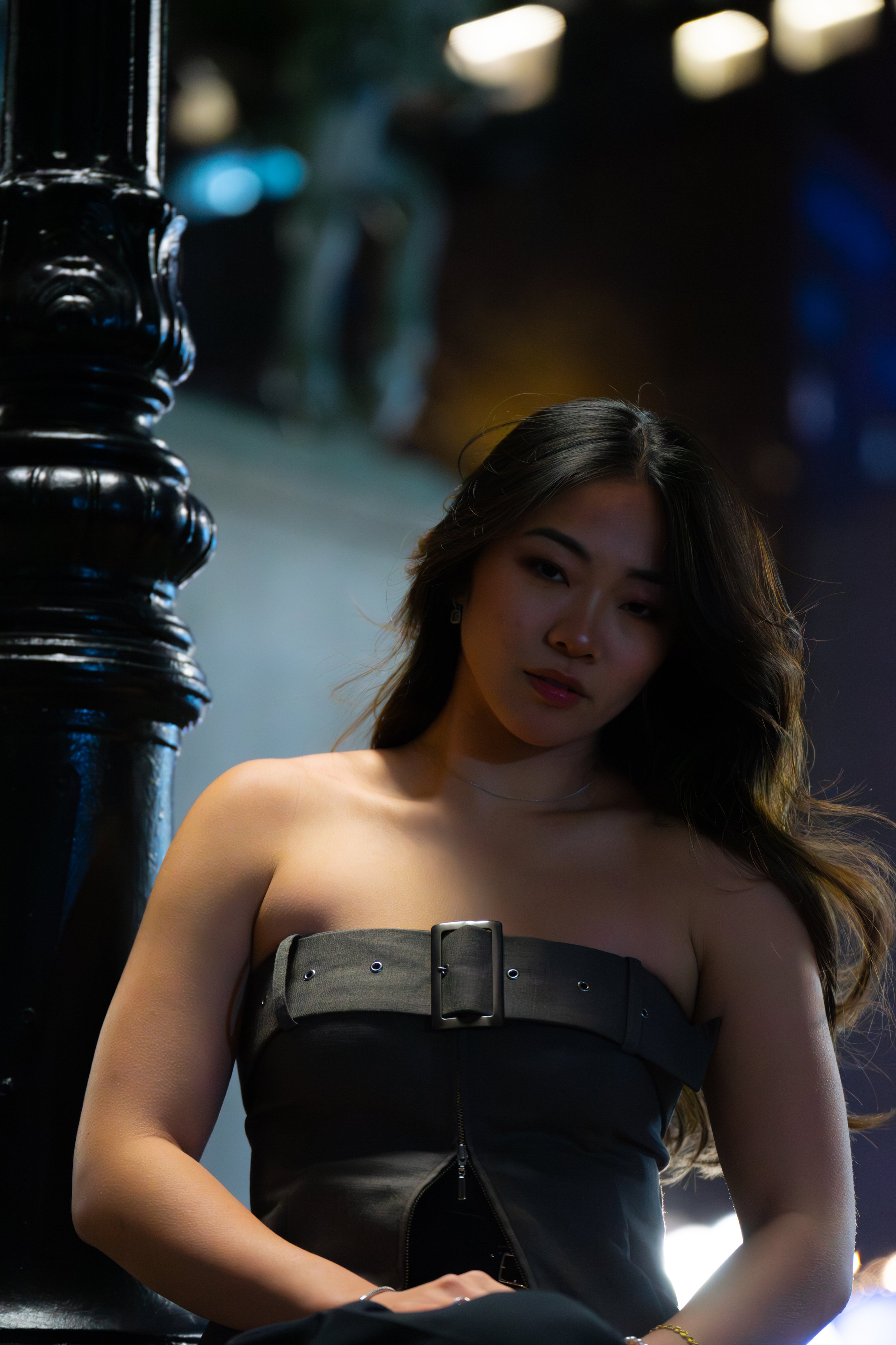 A young woman with long dark hair, wearing a strapless black top with a buckle, standing next to a black pole in an outdoor setting at night.