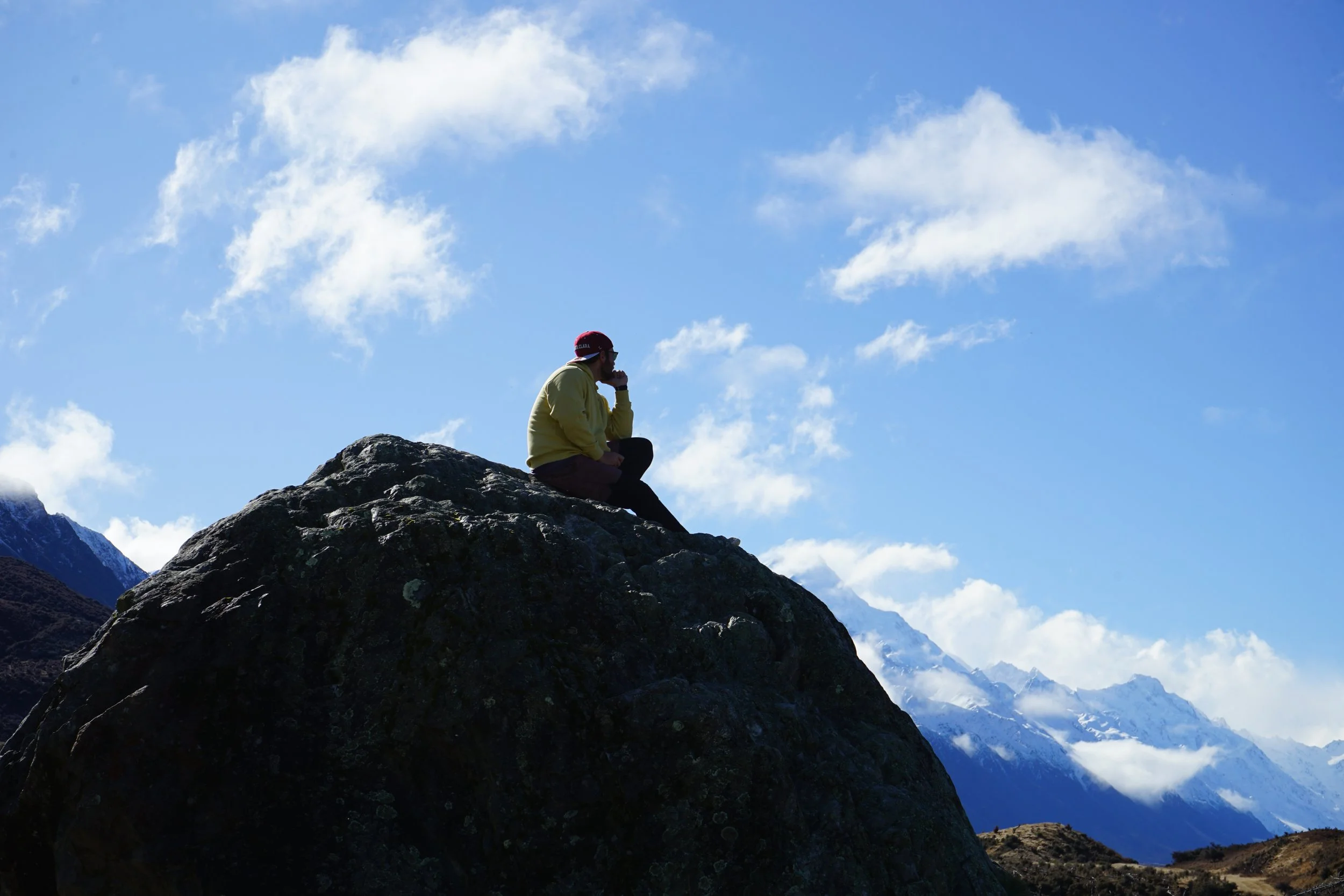 A person sitting on a large rock formation, looking at snow-capped mountains and a blue sky with scattered clouds in the background.