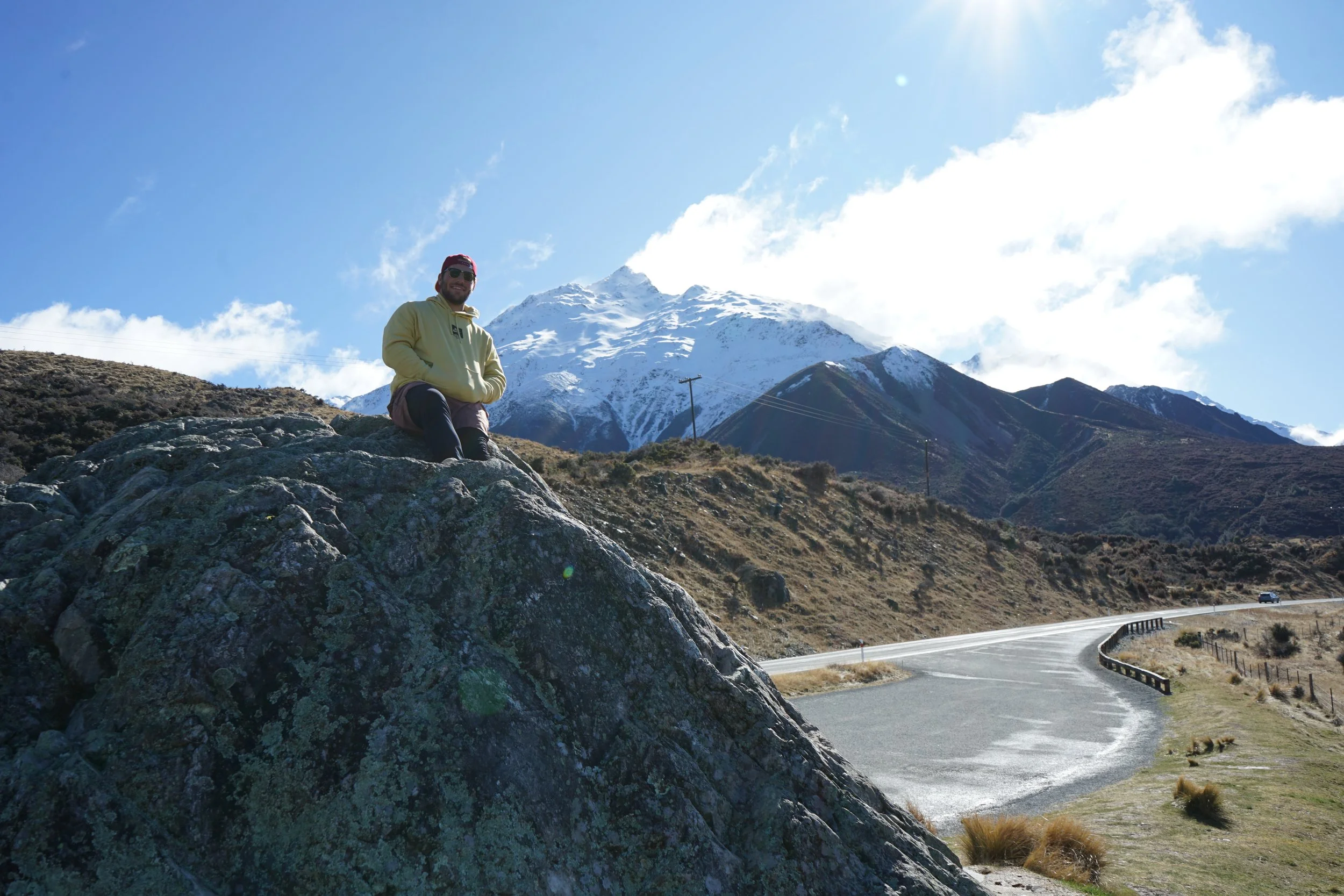 A man wearing a yellow hoodie, red cap, and sunglasses, kneeling on a large rock with a snow-capped mountain in the background, alongside a winding mountain road.
