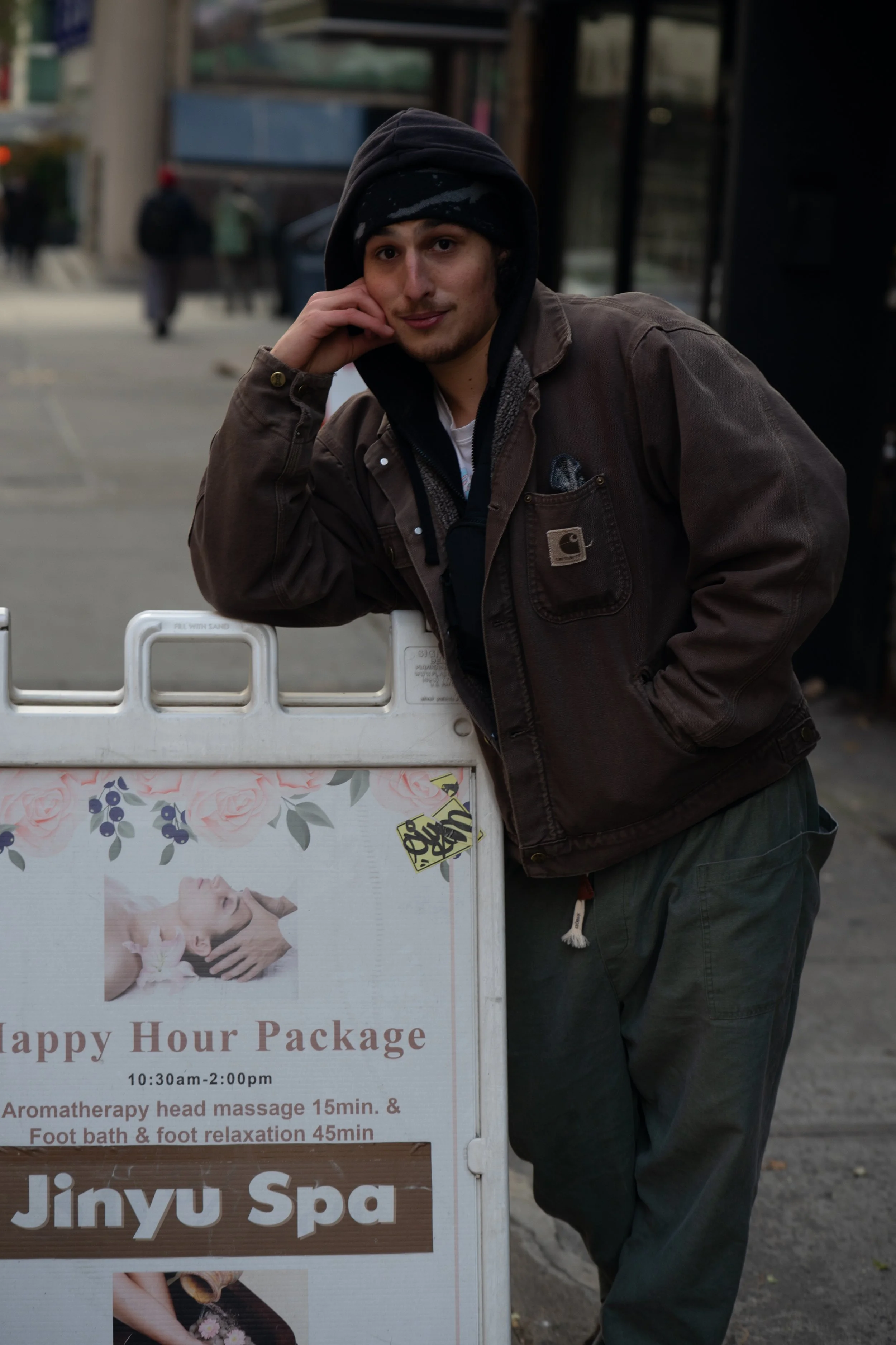 A man leaning on a sidewalk sign outside a spa, wearing a brown jacket and black hoodie, with hands in pockets.