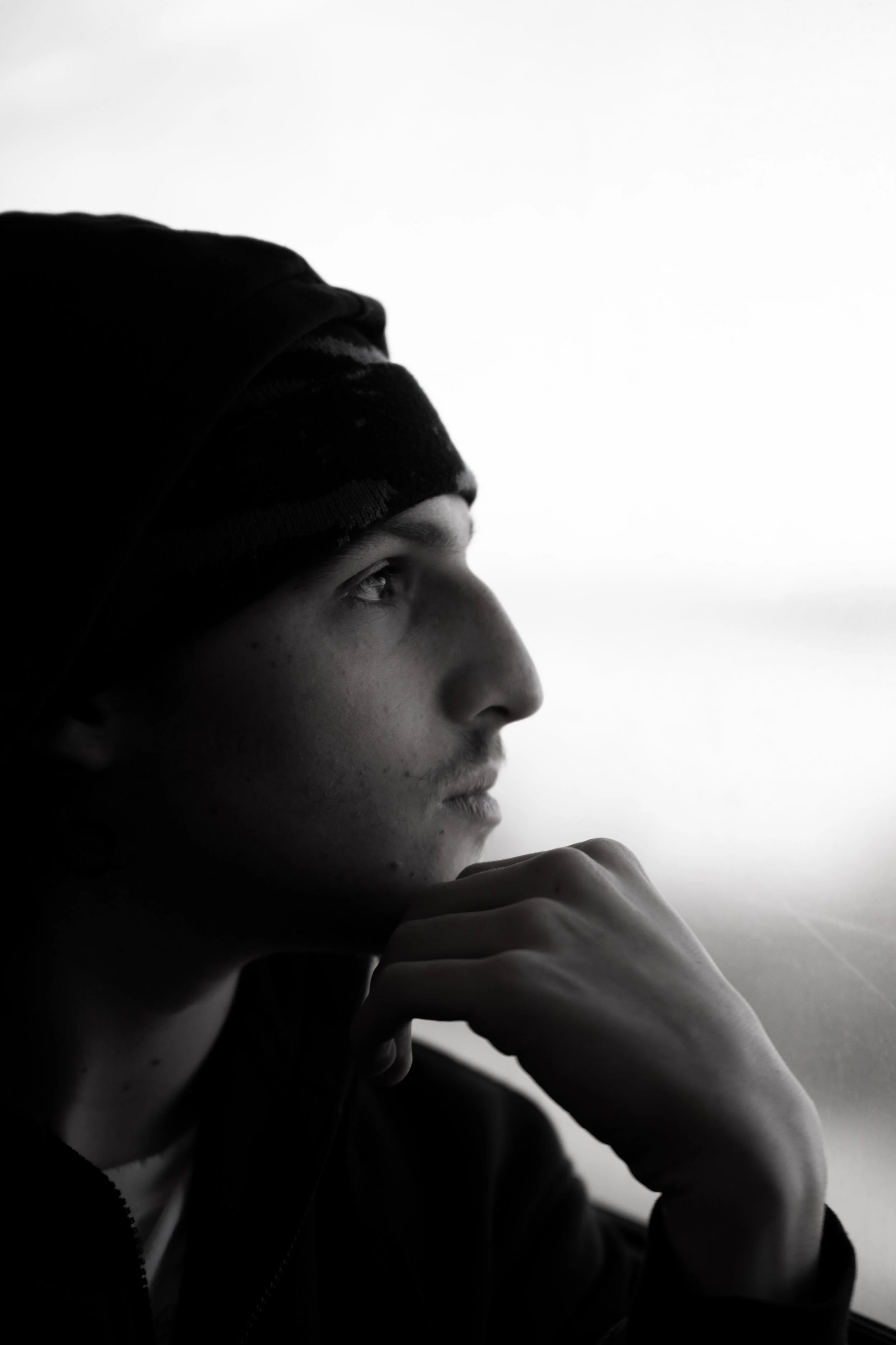 A black and white photo of a young man gazing thoughtfully out of a window, with his face resting on his hand.