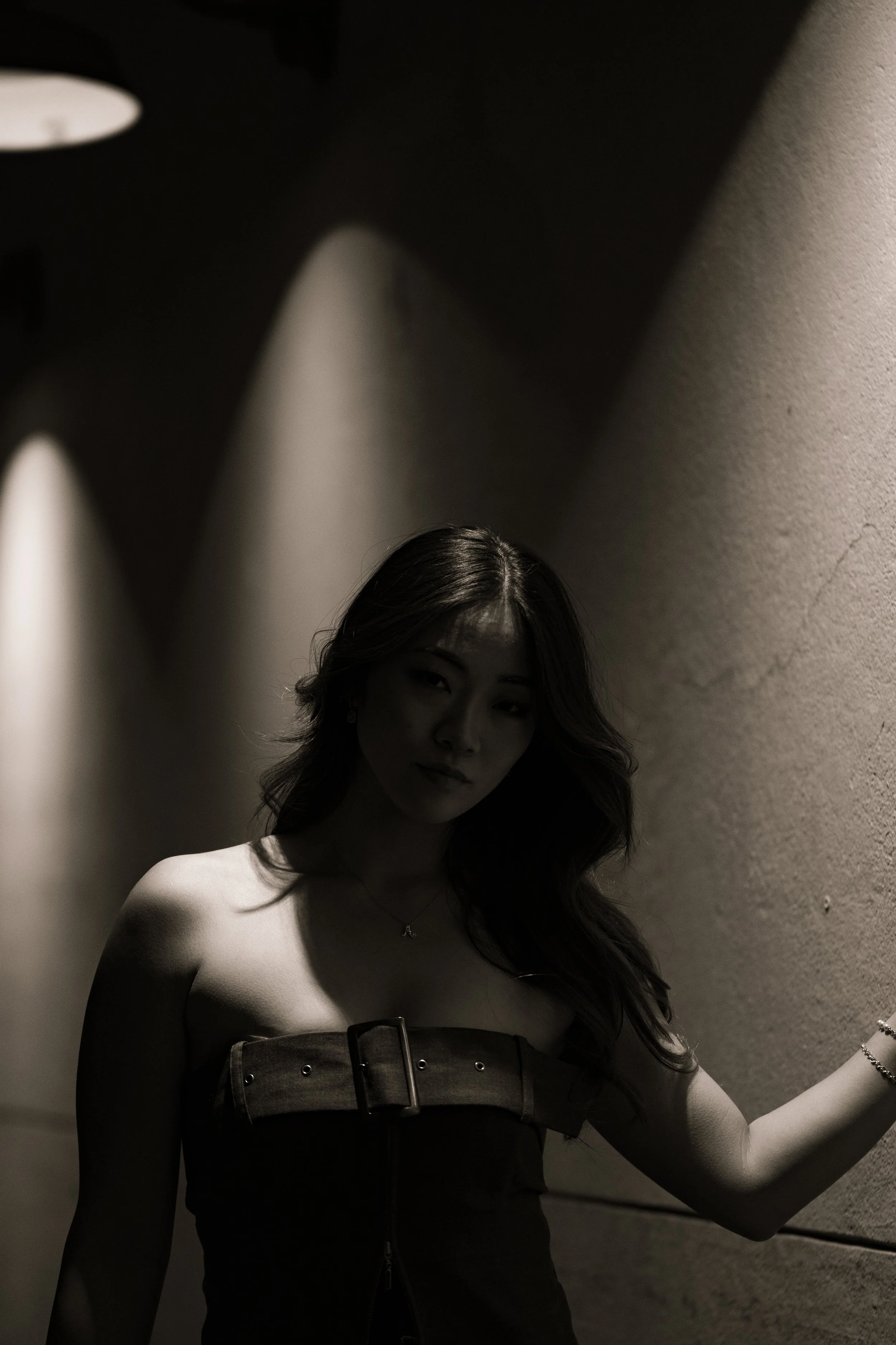 A black and white photo of a woman with long wavy hair, wearing a strapless top, standing against a textured wall with two wall lights creating a shadowy effect.