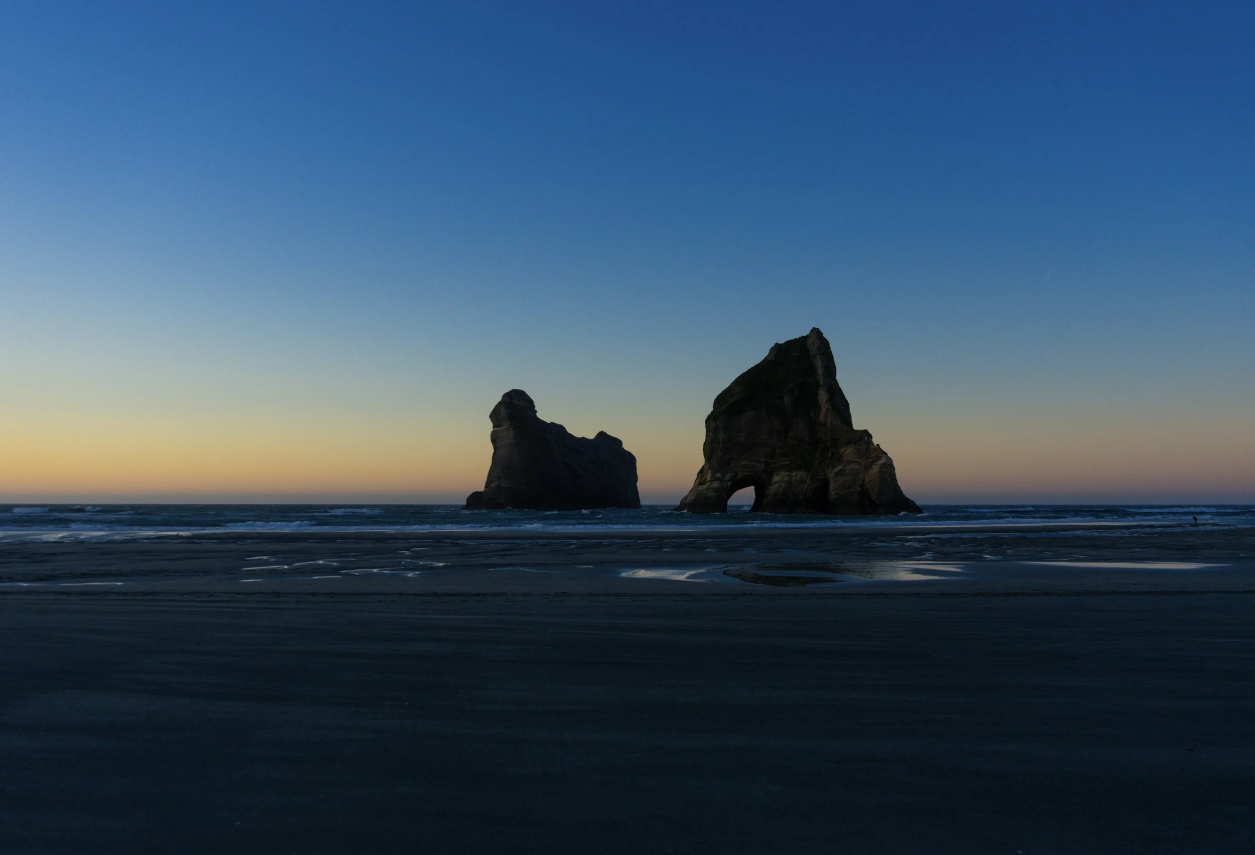 Sunset at the beach with two large sea stacks, one with a hole at the base, on dark sand with wet patches and gentle waves.