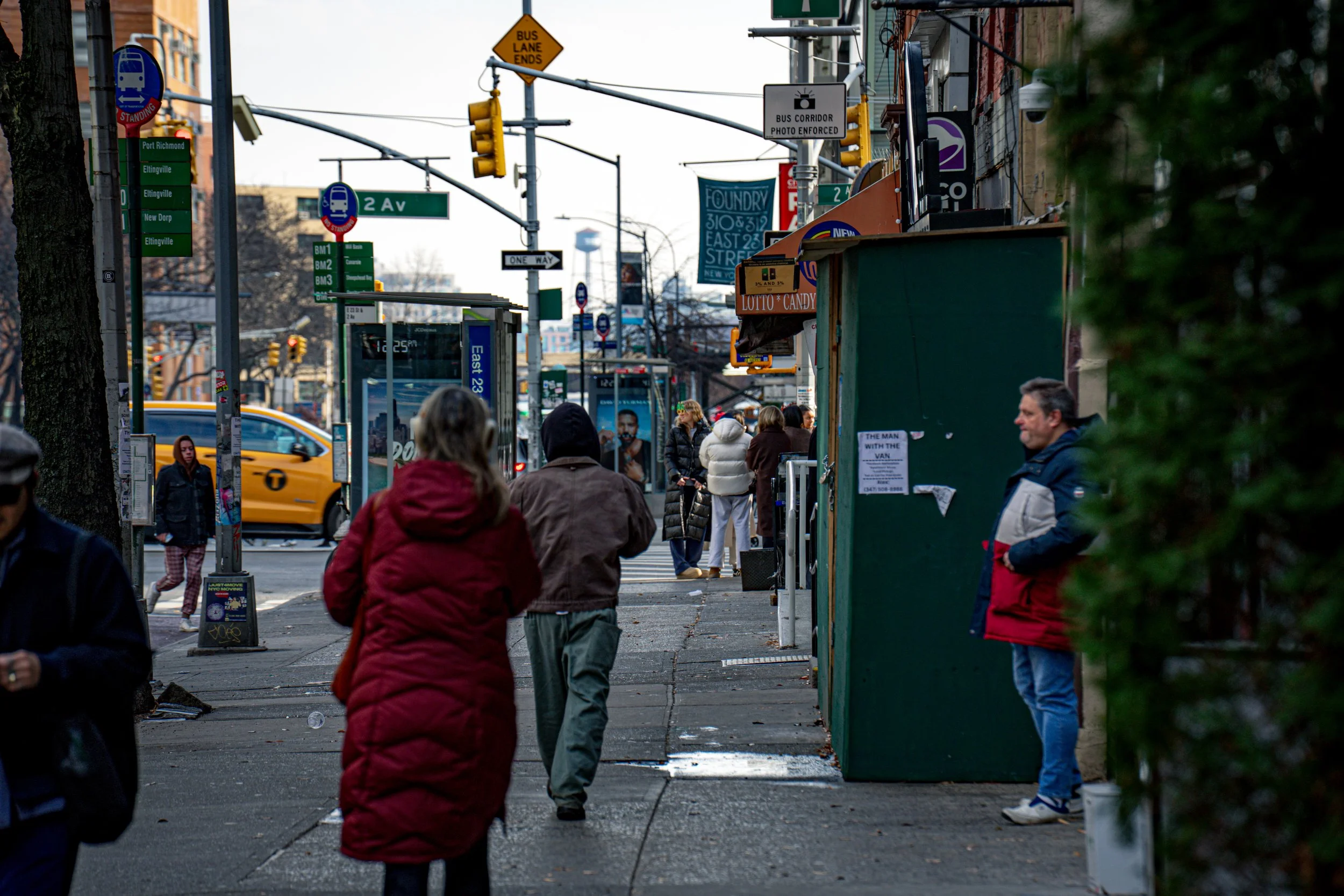 People waiting at a city bus stop on a sidewalk, with a yellow taxi driving by. There are various street signs, traffic lights, and storefronts visible in the background.