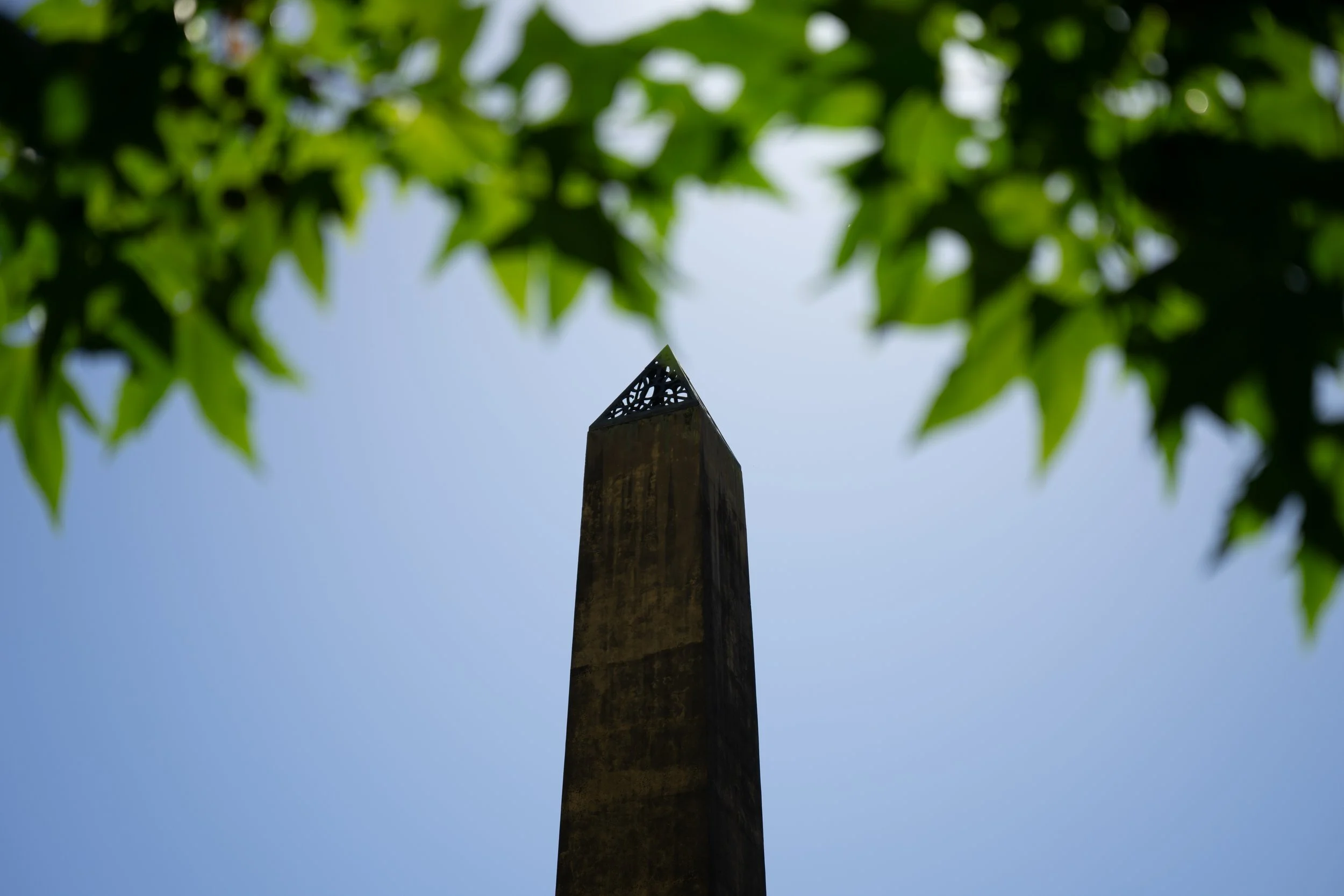 Upward view of a tall, dark stone obelisk with decorative top, framed by green tree leaves against a blue sky.
