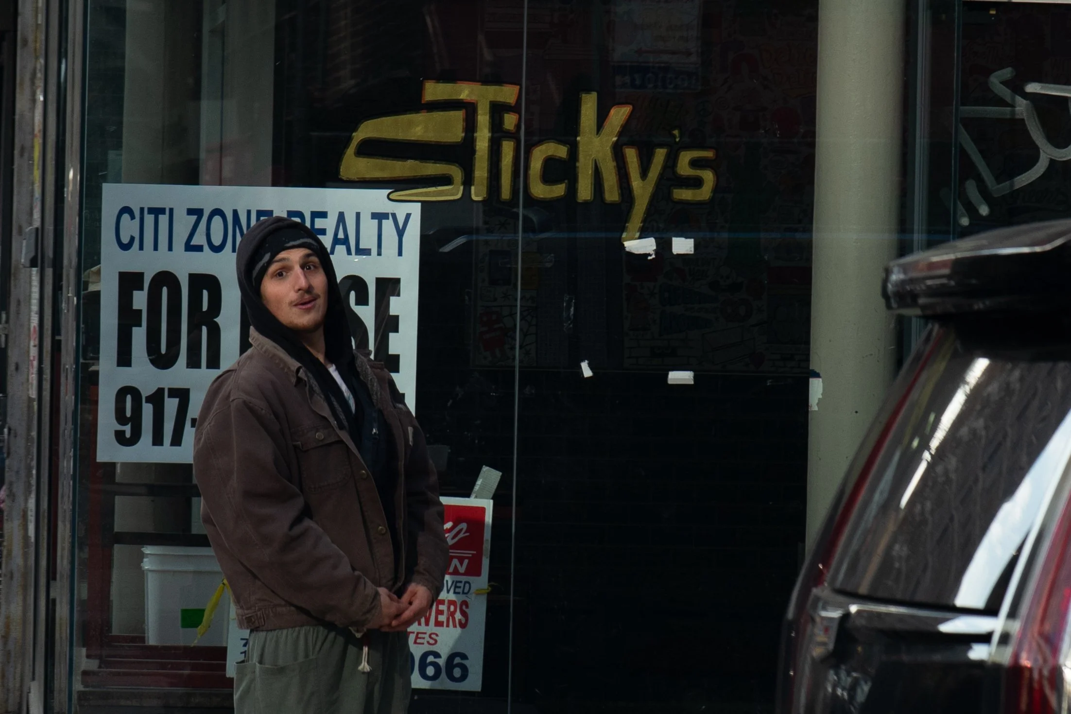 Young man in a brown jacket, black hoodie, and beige pants standing outside a store with a sign reading 'Sticky's.' There are also 'For Lease' and 'Citi Zone Realty' signs in the window.