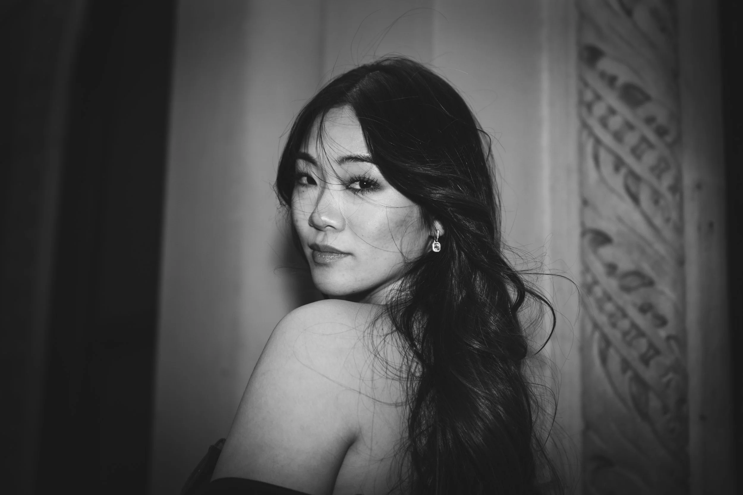 A black and white portrait of a woman with long dark hair, looking over her shoulder with a slight smile, earrings, and a bare shoulder, standing near a carved wall.